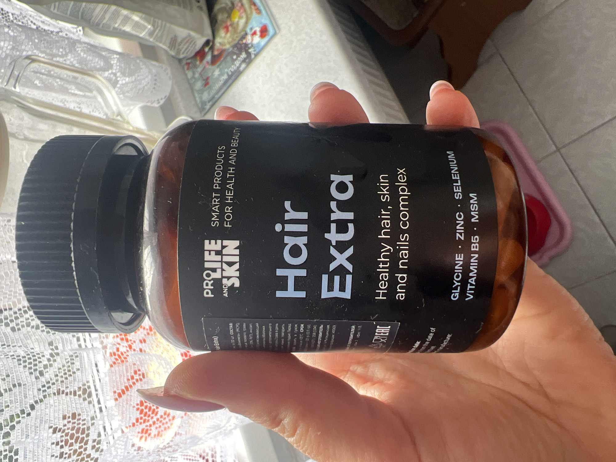 The image shows a hand holding a bottle of "Hair Extra" supplement. The bottle is amber-colored with a black cap and a black label. The label displays the brand "PROLIFE AND SKIN," the product name "Hair Extra," and a description "Healthy hair, skin and nails complex." Below this, it lists ingredients such as "GLYCINE, ZINC, SELENIUM, VITAMIN B5, MSM." The background appears to be an indoor setting with a window covered by a white lace curtain. The image does not provide any information about the location in Saint Petersburg, Russia.