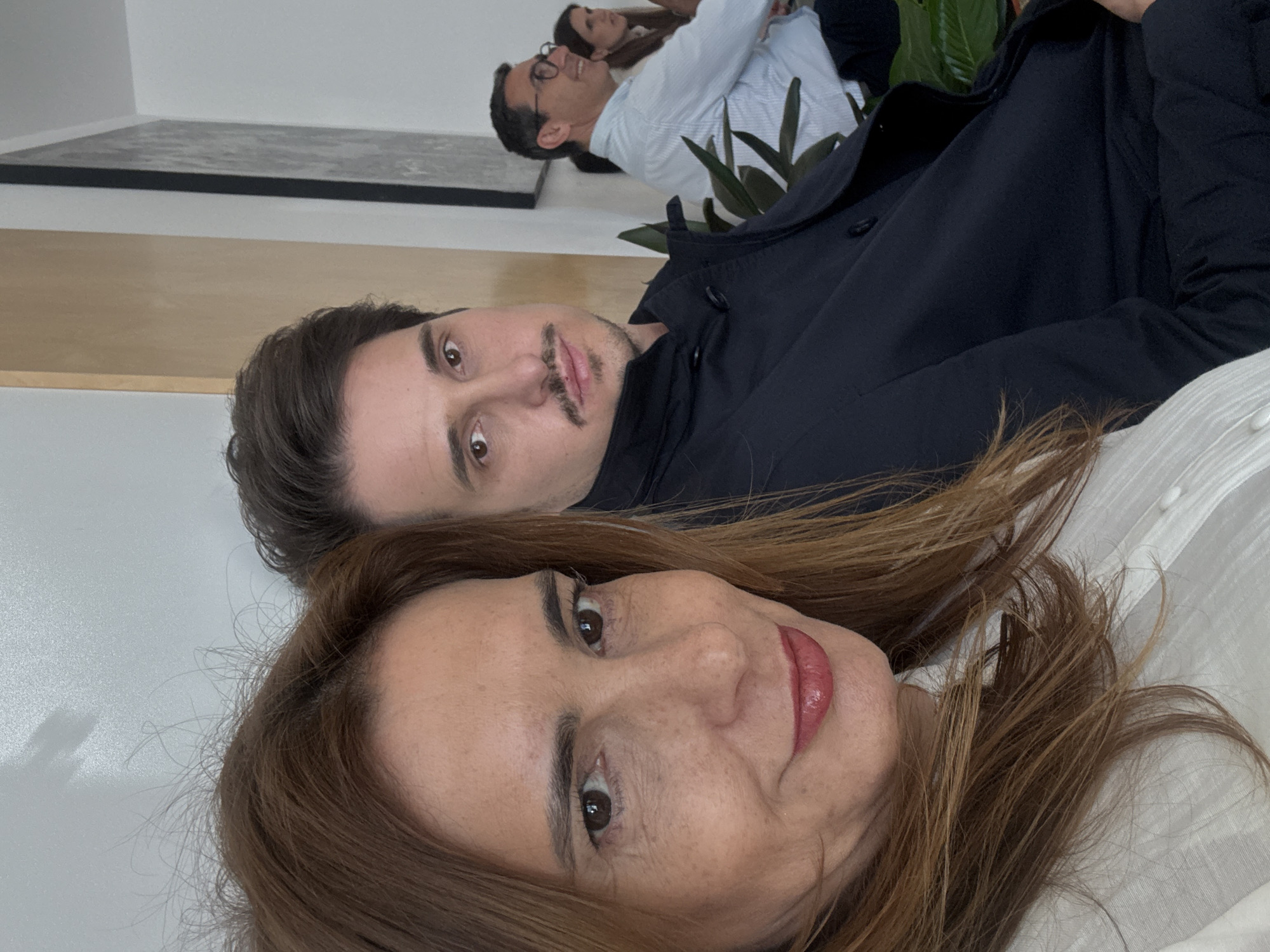 Three adults are visible in an indoor setting. The foreground features a woman with light brown hair and fair skin, wearing a white shirt. Behind her, a man with dark hair and a mustache is wearing a dark blue jacket. In the background, partially obscured by a potted plant, is another man with glasses wearing a light blue and white striped shirt, next to a woman. A grey and black artwork is visible on the wall behind them. The location is unknown, the city is also not discernible.