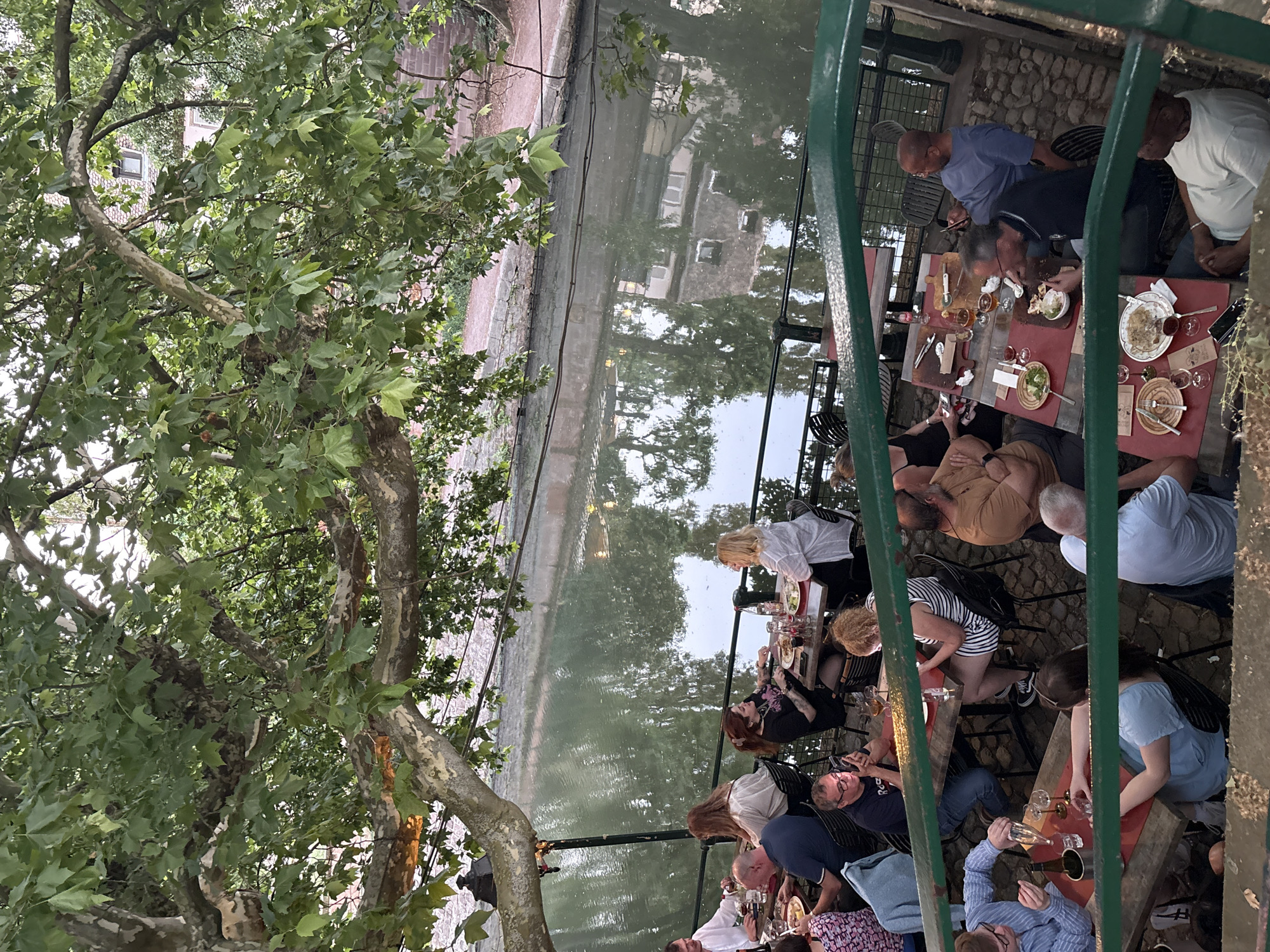 The image shows an outdoor dining scene in Strasbourg, France. People are seated at multiple red tables along a canal, enjoying food and drinks. A large tree with green leaves is visible on the left side, partially obstructing the view. The canal reflects buildings and the surrounding environment. A green metal railing is present in the foreground. The atmosphere appears casual and convivial.