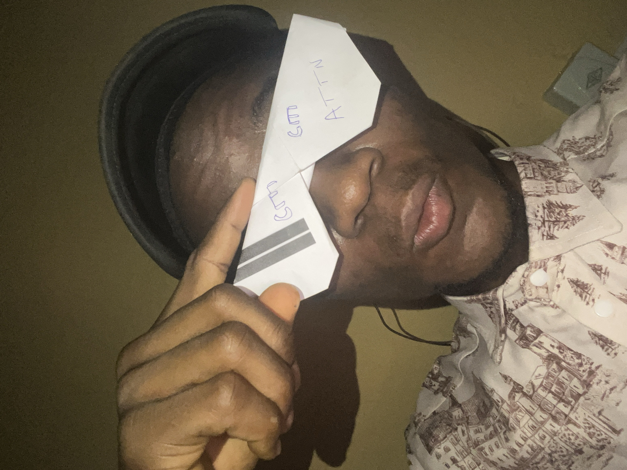 A man is holding a paper airplane over his eyes. "ATTN" and "GEM" are written on the airplane. He is wearing a hat and a patterned button-down shirt. The location is Minna, Nigeria.