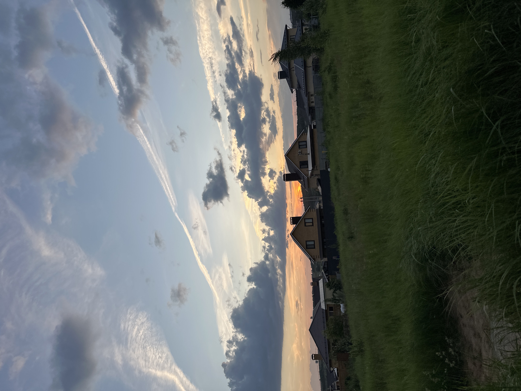 The image depicts a suburban landscape at dusk in the city of Estachi, Russia. In the foreground, a field of tall, green grass sways gently. Behind the field, several houses with tiled roofs and visible chimneys are lined up, suggesting a residential area. The sky is a gradient of blue and orange, with scattered clouds and visible contrails from aircraft. The overall impression is a peaceful, rural setting transitioning from day to night.