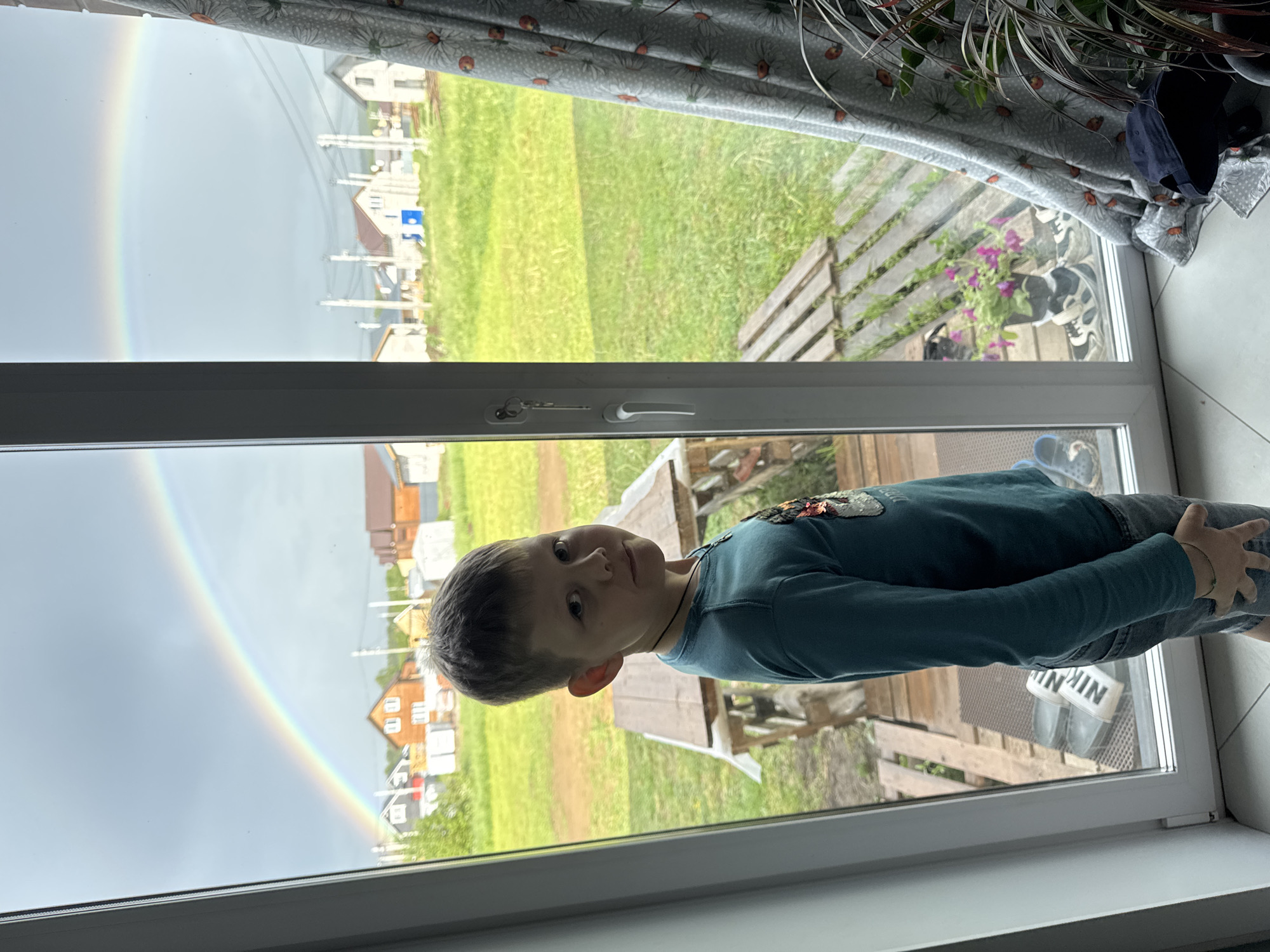 A waist-up, full shot of a young boy looking out of a window reveals a double rainbow arcing above a residential area. The boy, positioned to the right of the frame, is wearing a long-sleeved turquoise shirt, grey shorts, and sandals. The window offers a view of a green lawn, a wooden patio or deck, houses in the background, and a visible rainbow on the left side. The curtain and a plant partially frame the top of the window. Location: Ферма, Russia.