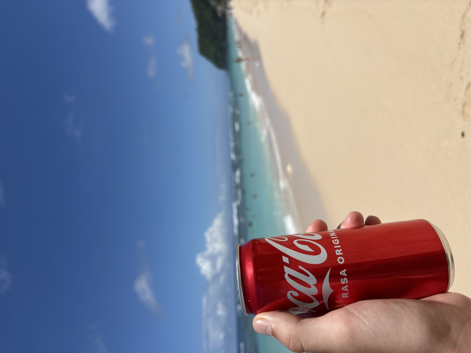 A photograph captures a person's hand holding a red Coca-Cola can with white lettering, displaying the words "Coca-Cola" and "RASA ORIGINAL." The backdrop shows a beach scene with light sand, turquoise water, and a clear blue sky dotted with white clouds. In the distance, a densely vegetated cliff or headland can be seen. People appear to be enjoying the water and beach. Given the details, the location is likely Ungasan, Indonesia.