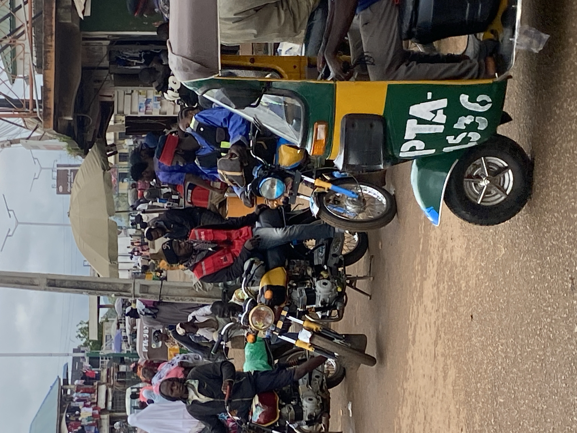 This image captures a bustling street scene in Bukuru, Nigeria, with a focus on the local modes of transportation and commerce. In the foreground, a green and yellow auto-rickshaw, identified by the registration number "PTA-1536" painted in white on its side, is parked. Beside it, a yellow and black motorcycle is also present.
Several individuals are visible, engaged in various activities. A man wearing a red vest and black cap is seated on a motorcycle. Another man in a blue uniform and a red band on his head is also on a motorcycle, holding a light brown bag. Behind them, a woman in a white headscarf and a woman in a pink headscarf are walking. In the background, a market area is evident with stalls displaying clothing and other goods, and more people are milling about. The environment suggests a lively, urban setting with some overcast sky overhead.