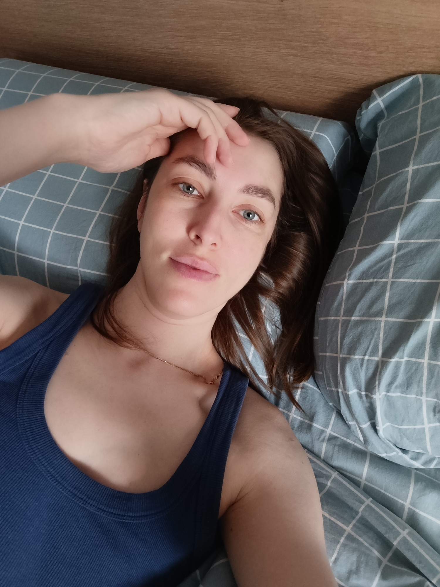 A young woman lies in bed, her head resting on a pillow with a light blue and white checkered pattern. She wears a dark blue tank top and a gold necklace. Her brown hair is slightly curled, framing her face. She is looking directly at the camera, her right hand resting on her forehead. The headboard of the bed is visible in the upper right corner of the image. The location is presumed to be Chaikovsky, Russia.