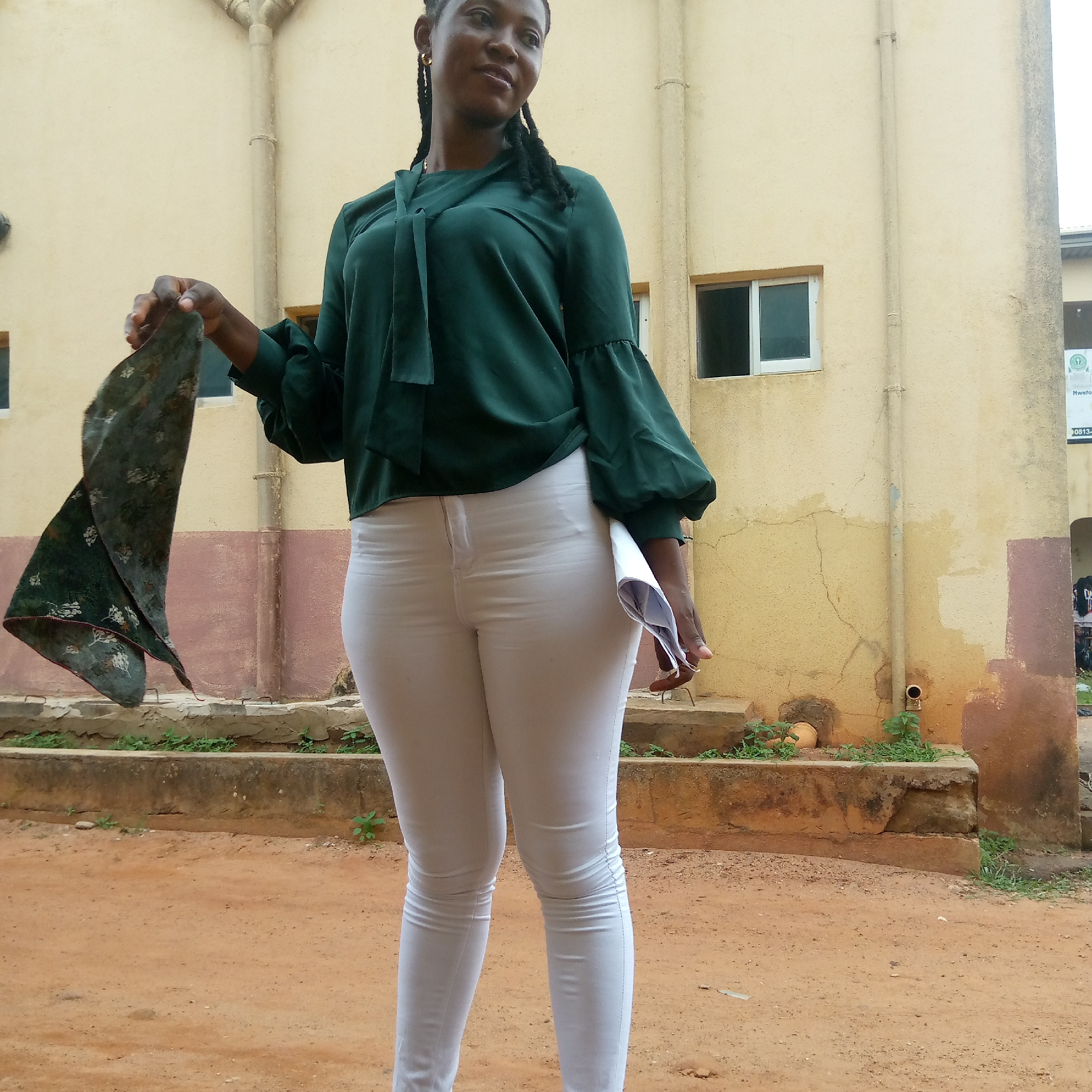 A woman stands in front of a building, possibly in Ukwu-Abwa, Nigeria. She wears a green blouse with a tie at the neck and white pants. She holds a scarf in one hand and papers or a mask in the other. The building behind her has a beige exterior and some weathering. The ground is dirt.