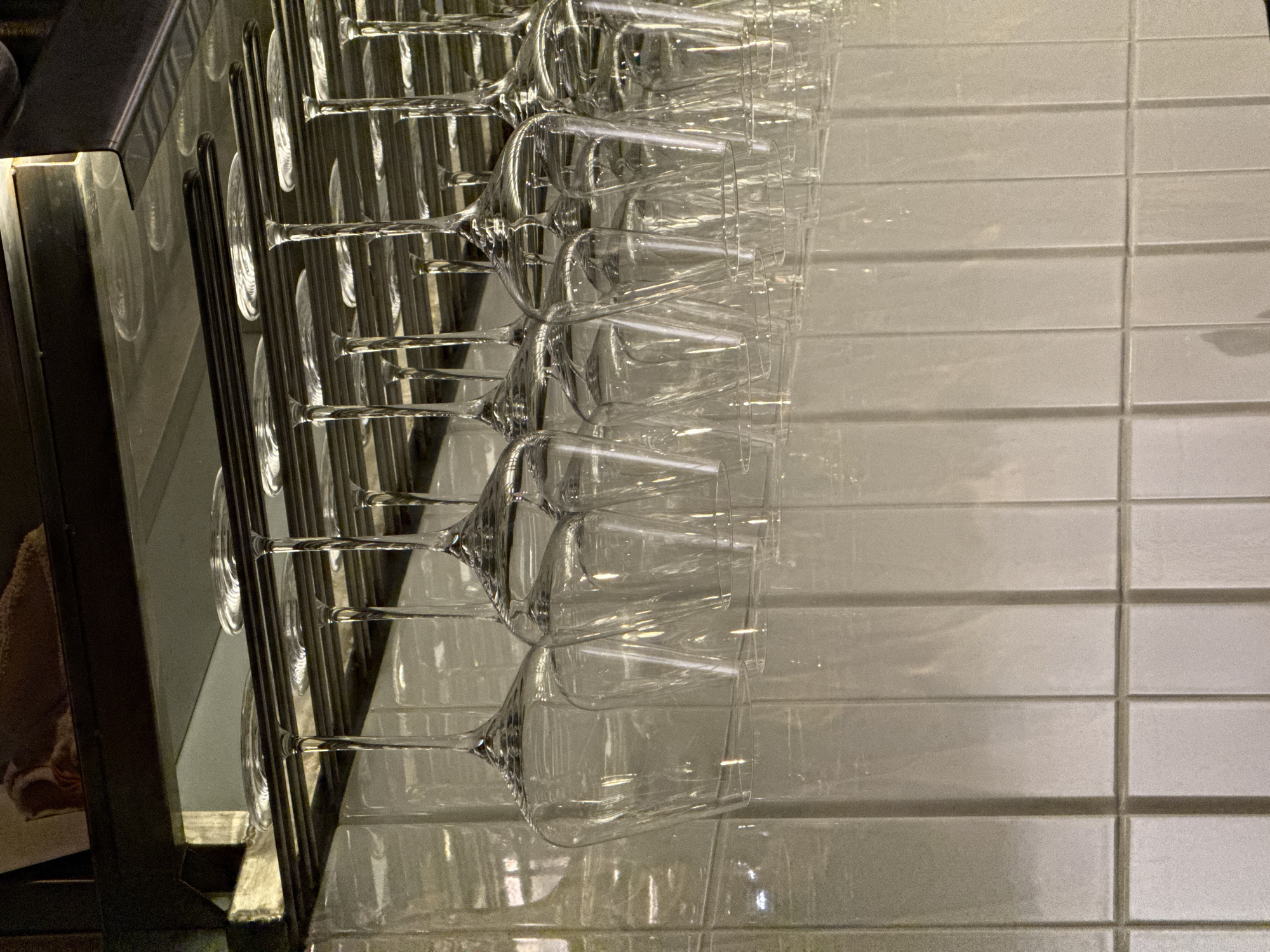 The image captures a precisely arranged row of clean, clear stemmed glasses, likely wine or cocktail glasses, hanging upside down from a sleek metallic rack. These glasses are uniformly spaced and appear identical in shape, suggesting a prepared or storage area in a commercial establishment such as a bar, restaurant, or hotel.
In the background, a light-colored tiled wall features a grid pattern of rectangular tiles, possibly subway tiles, which exhibit a subtle sheen and reflect some of the ambient light. To the far left, a darker structure, which could be part of a counter or cabinet made of dark wood or metal, frames the scene. A bright reflection or light source illuminates the edge of this structure.
The lighting is artificial, creating soft reflections and subtle variations in brightness across the transparent surfaces of the glasses and the polished tiles. There is no explicit activity taking place; the scene conveys a sense of orderliness, cleanliness, and readiness for service within an indoor environment. No text is visible in the image.