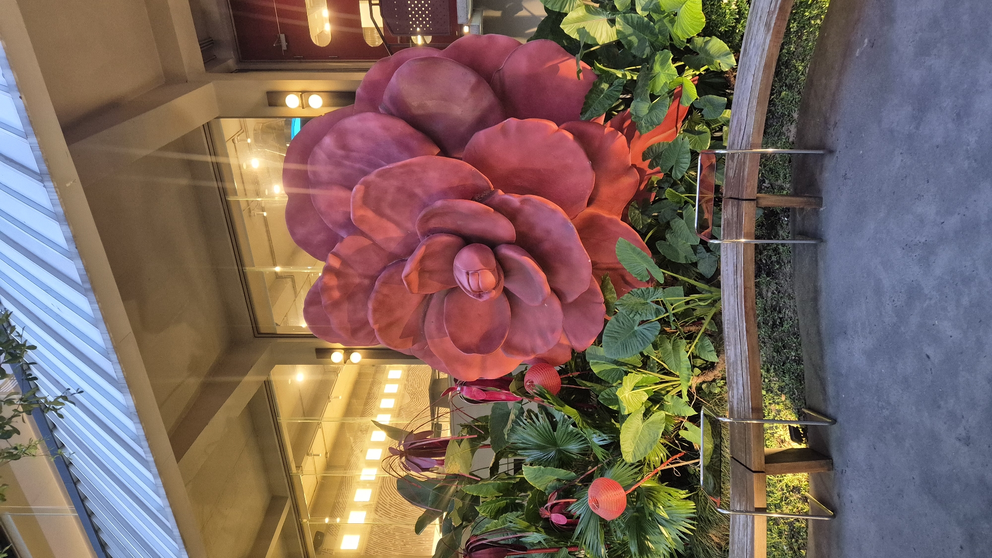 The image shows a large, artificial, deep red flower sculpture as the central subject, surrounded by lush green foliage and some red decorative lanterns. In the foreground, a curved wooden bench with metal supports sits on a grey concrete pathway. Behind the foliage and sculpture is a building with large glass windows, through which the warm glow of interior lighting is visible. The building has a modern architectural style with exposed concrete beams and a corrugated metal roof. The scene appears to be an outdoor seating area, possibly at a restaurant, cafe, or public space. The overall impression is one of a decorative and inviting outdoor setting. This scene is located at the Da Nang Museum of Cham Sculpture in Da Nang, Vietnam.