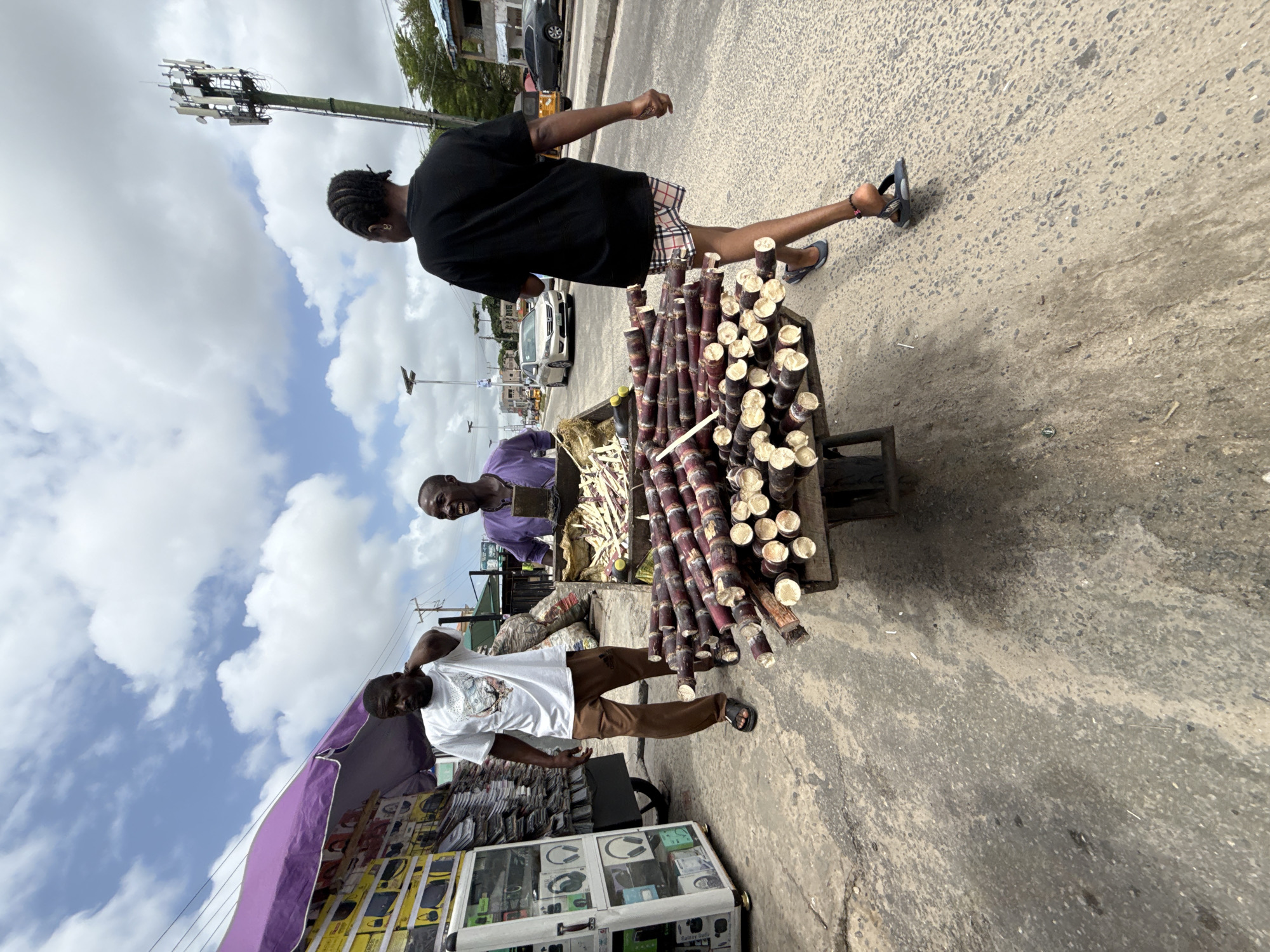 The image depicts a street scene in Ado, Nigeria. A street vendor is selling sugar cane from a pushcart, with bundles of the purple stalks visible. A second person, likely the vendor's partner or assistant, stands beside the cart. A pedestrian wearing a black t-shirt and plaid shorts is walking past the cart. Another street vendor is seen selling electronics and accessories from a display case under a purple awning. In the background, cars are parked along the side of the road, and a telecommunication tower is visible. The setting is a busy urban environment with pedestrians and street vendors.