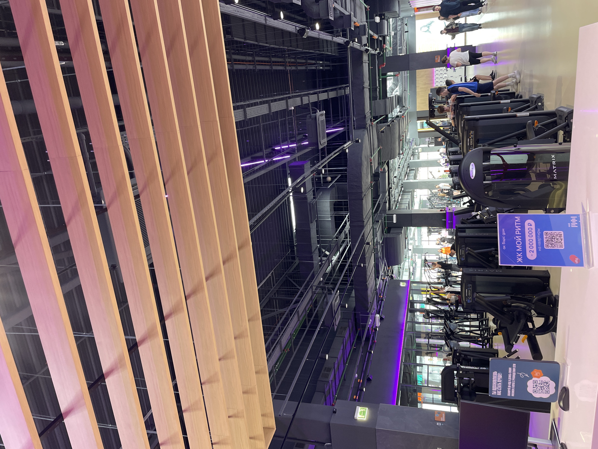 The image shows the interior of a modern gym in Kazan, Russia. In the foreground and to the left are vertical, parallel wooden slats, seemingly part of a design feature or wall panel. The ceiling is visible, with a complex network of metal beams, pipes, and ductwork, some accented with purple lighting. The gym itself is well-equipped with various Matrix-branded exercise machines. People are seen working out on the equipment, including chest press and leg curl machines. Several signs are in place, one reads "ЖК МОЙ РИТМ - 2000 000 Р на квартиры", advertising an apartment complex for 2,000,000 rubles. Another sign has a QR code, inviting members to share their opinions with gym management. The gym appears to be spacious and well-lit.
