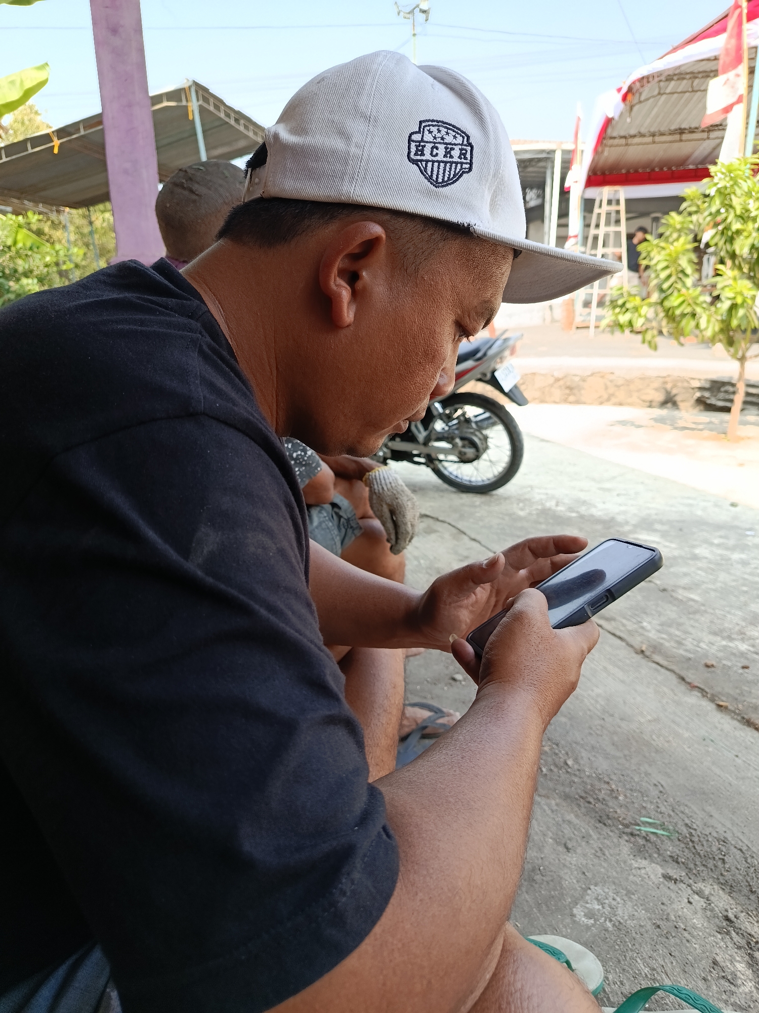 A man wearing a black t-shirt and a white baseball cap with "HCKR" embroidered on it is holding a black smartphone and looking at it. He appears to be sitting outside, as evidenced by the concrete ground and a motorcycle in the background. The setting looks like it could be Jurug, Indonesia, with a tiled roof structure and what appear to be Indonesian flags in the background, suggesting a possible public event or gathering. Sunlight is casting shadows, indicating it's daytime. There is another person partially visible in the background, seated on the ground.