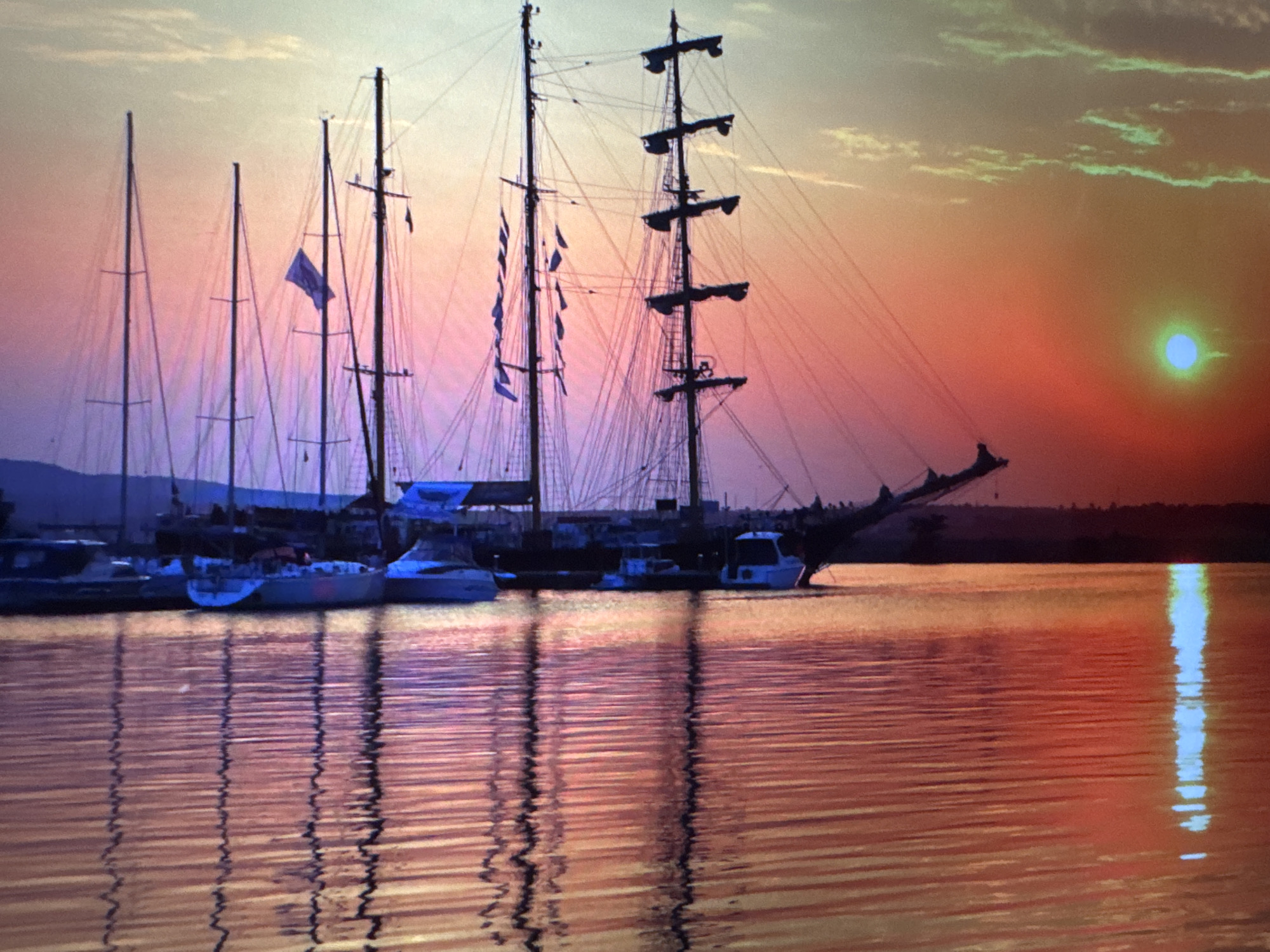 The image captures a serene sunset scene at a harbor or marina, featuring several boats silhouetted against a vibrant sky.
In the foreground, a tranquil body of water reflects the intense colors of the setting sun, creating rippling patterns of orange, red, and purple. Numerous boats are docked or anchored, their dark forms contrasting with the brightly lit horizon. The most prominent vessels are a large multi-masted schooner or tall ship, and several sailboats of varying sizes. The tall ship stands out with its intricate rigging and multiple yards, along with several small pennants flying from its lines. Another sailboat closer to the center displays a larger, dark blue or purple flag. Smaller yachts and motorboats are also visible along the dock line.
The sky is a dramatic display of sunset hues, transitioning from a deep, fiery orange and red near the horizon to softer purples and light blues higher up. Wispy clouds are scattered across the sky, catching the light and appearing in shades of light green and orange. The sun itself is a striking, distinct disk of bright blue-green, positioned low in the sky on the right side of the frame, casting a shimmering vertical reflection on the water. A dark, indistinct landmass or shoreline forms the distant background behind the boats.
The overall atmosphere is peaceful and picturesque, highlighting the beauty of the twilight hour. No visible text is present in the image.