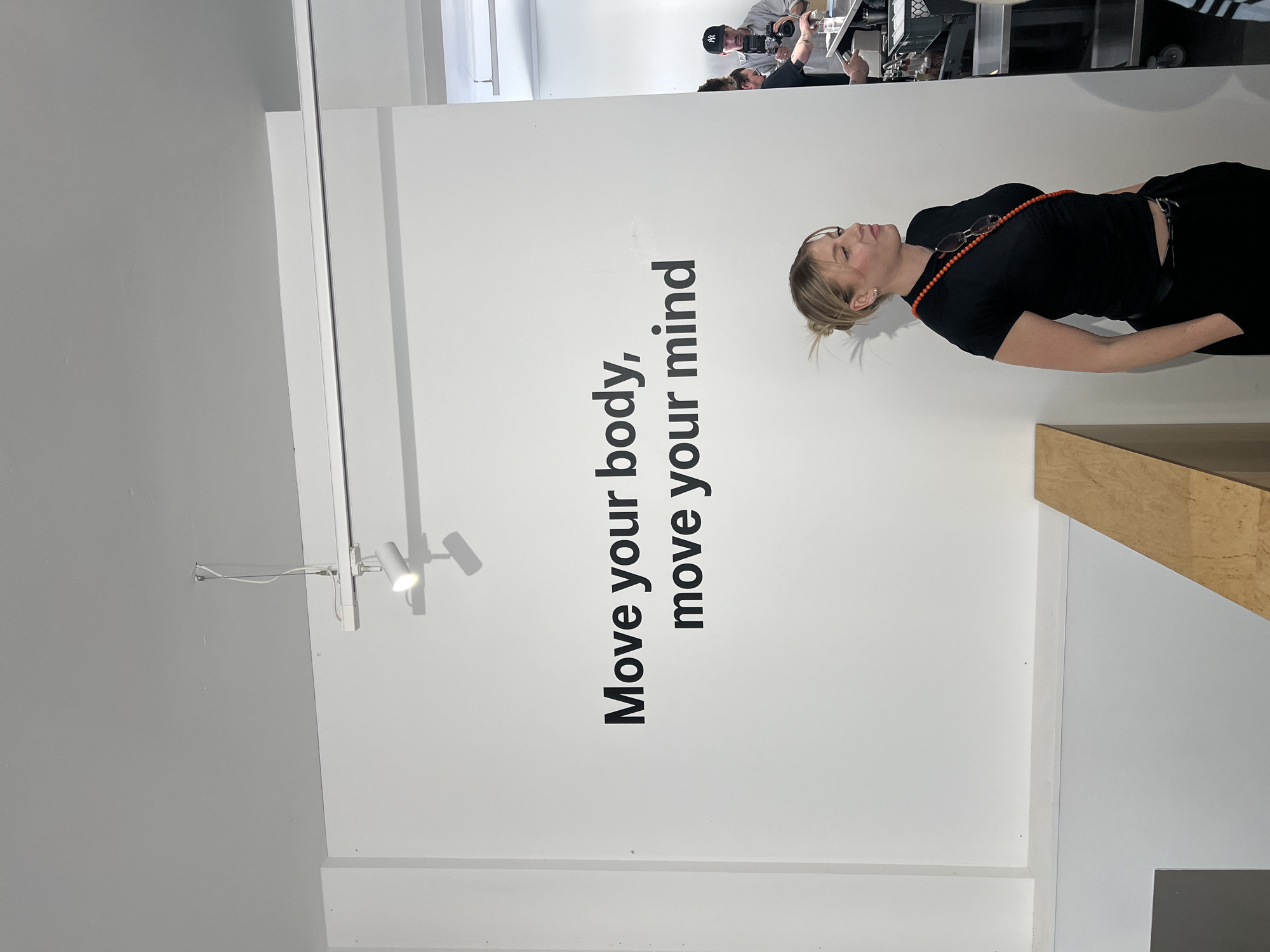 A brightly lit indoor scene, likely a gallery, studio, or event space, features a woman standing in front of a large white wall with prominent black text. The woman, with blonde hair pulled back in a bun, wears a black t-shirt, black trousers, and an orange beaded necklace from which sunglasses hang. She is seen in profile, looking upwards and to her right, with a serene or contemplative expression.
The large black sans-serif text on the white wall reads: "Move your body, move your mind".
In the background, positioned further away and behind a counter-like structure, several individuals are visible. One person, wearing a New York Yankees baseball cap, is holding a camera or similar recording equipment, appearing to film or photograph the scene. Other people are partially visible around them, suggesting a professional media setup or an event in progress.
The space is minimalist with white walls and a high ceiling from which track lights are suspended. A light grey wall is visible in the background on the left. In the lower right foreground, a part of a light brown wooden structure, possibly a plinth or display stand, is visible. The bright, even lighting suggests daytime or a well-lit interior. The overall atmosphere suggests a contemporary art exhibition, promotional event, or fitness-related display in London, United Kingdom.