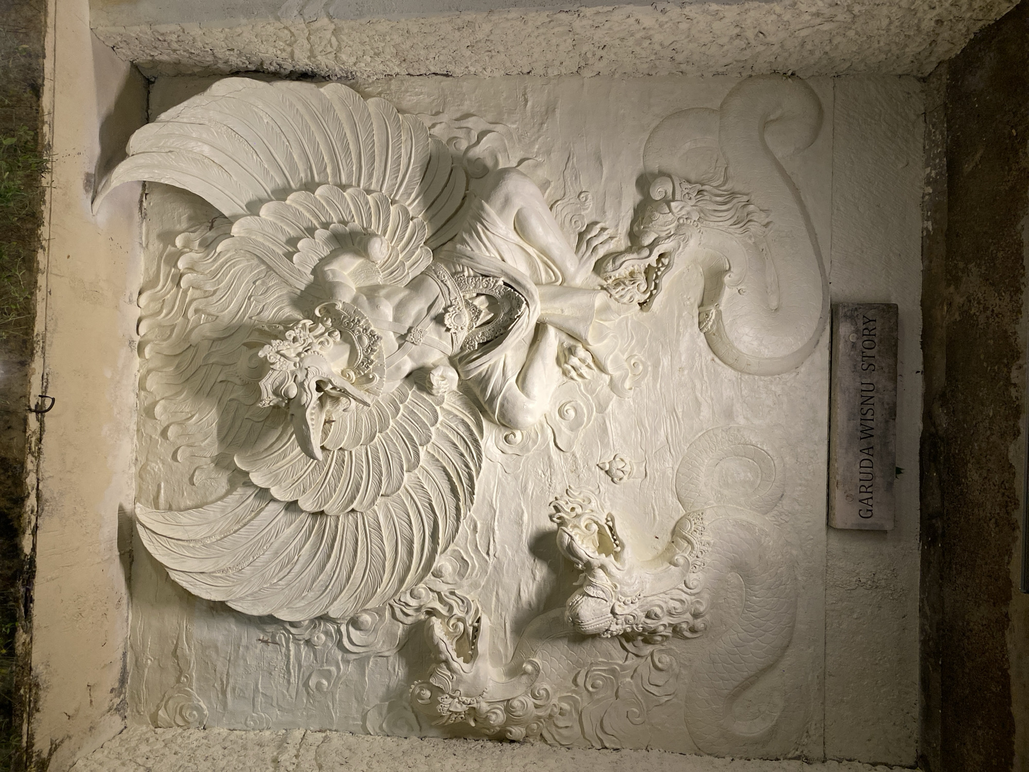 The image shows a white, low-relief stone carving of Garuda, a mythical bird-like creature, with large spread wings, sitting in a meditative pose. Garuda is depicted with a bird's head and talons, but a human-like torso and legs. It is adorned with ornate jewelry and a flowing garment. Flanking Garuda are two serpent-like creatures, also rendered in the same white stone, with detailed scales and fierce expressions. The background is textured to resemble clouds. Below the carving, a small rectangular stone plaque is inscribed with the words "GARUDA WISNU STORY." The carving is set within a recessed alcove with rough, textured stone walls on either side. The top of the alcove appears to be exposed to the elements, with some soil and greenery visible at the very top edge. The lighting suggests this is an outdoor sculpture, likely illuminated at night. This carving is located at Garuda Wisnu Kencana Cultural Park in Ungasan, Kuta Selatan, Bali, Indonesia.
