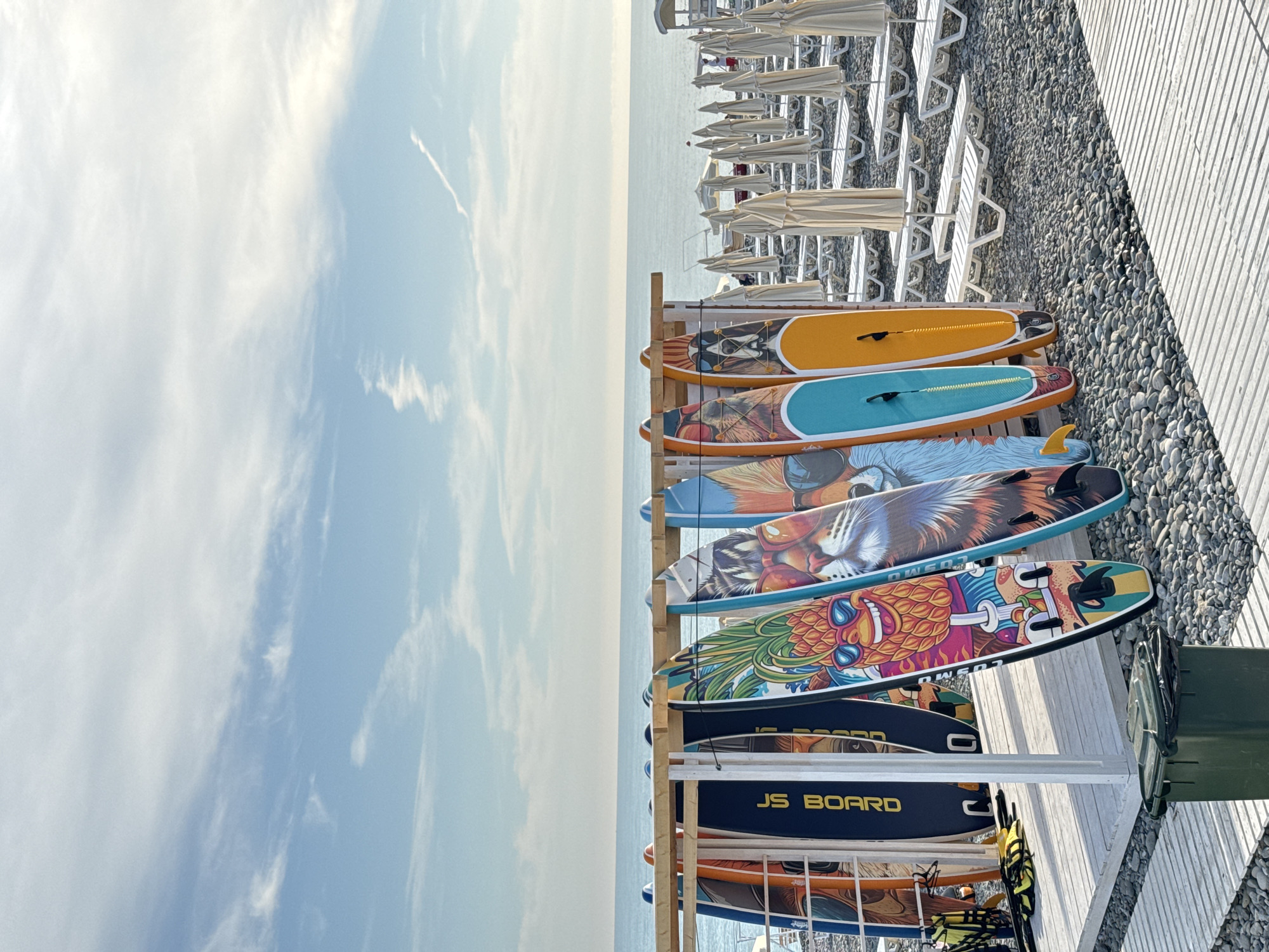 This image captures a sunny beach scene in Sochi, Russia, during daylight hours. In the foreground, a green lidded trash bin with a black liner is situated on a light-colored wooden slatted walkway. To the left, a white wooden rack holds approximately ten paddleboards (SUPs) upright. Distinct designs visible on the paddleboards include a dark blue board with "JS BOARD" in yellow text, a cartoon pineapple wearing sunglasses, a tiger face wearing sunglasses, a fox face wearing sunglasses, and a dog face wearing sunglasses on an orange-yellow board. Yellow life vests are stacked at the base of the paddleboard rack. The beach surface is composed of small, grey and tan pebbles. To the right, a large section of the beach is equipped with numerous rows of unoccupied white plastic lounge chairs, each accompanied by a folded, cream-colored beach umbrella. In the background, the calm, blue expanse of the Black Sea meets the horizon under a pale blue sky. The sky features scattered white, wispy cirrus clouds and a discernible white contrail from an aircraft in the upper right. Indistinct human figures are visible in the distance near the beach furniture on the far right.
