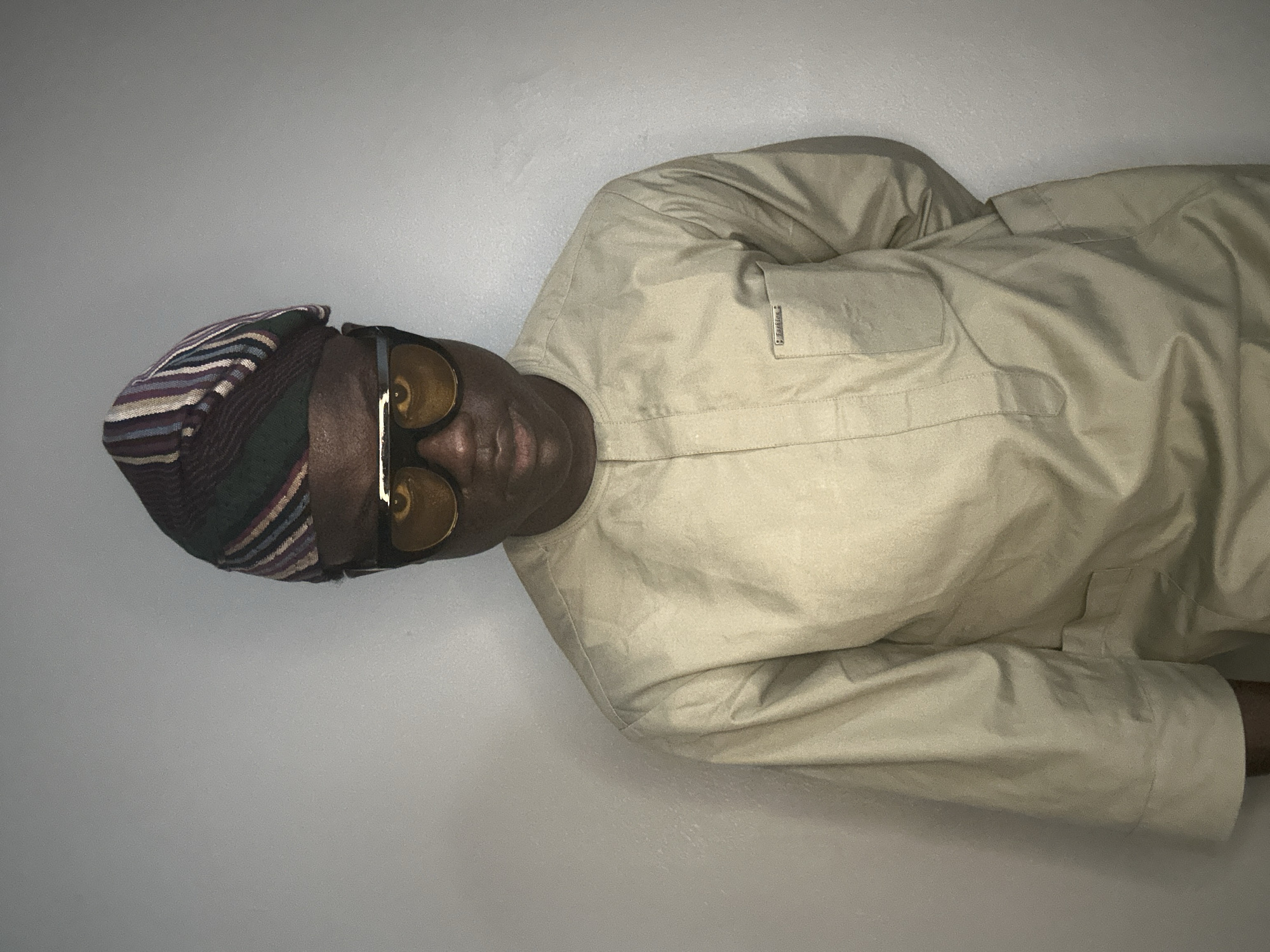 The image shows a person wearing a beige jacket and a colorful striped headwrap. The person is also wearing large, round sunglasses with yellow lenses. The person's face is dark-skinned and they are looking directly at the camera. The background is a plain, light-colored wall. The image is vertically oriented, and the person appears to be lying down or leaning back. There is a small tag on the jacket with text that is difficult to read. The overall impression is that of a posed portrait, with the subject displaying a confident and stylish demeanor. The lighting is somewhat flat, suggesting an indoor setting. The style of the headwrap might indicate a Nigerian origin, and the mention of Badagry, Nigeria as the location context supports this. There are no strong cues about the time of day or weather, but the indoor setting makes this information less relevant. The subject's expression is neutral, but the stylish attire and sunglasses suggest a sense of self-assurance.