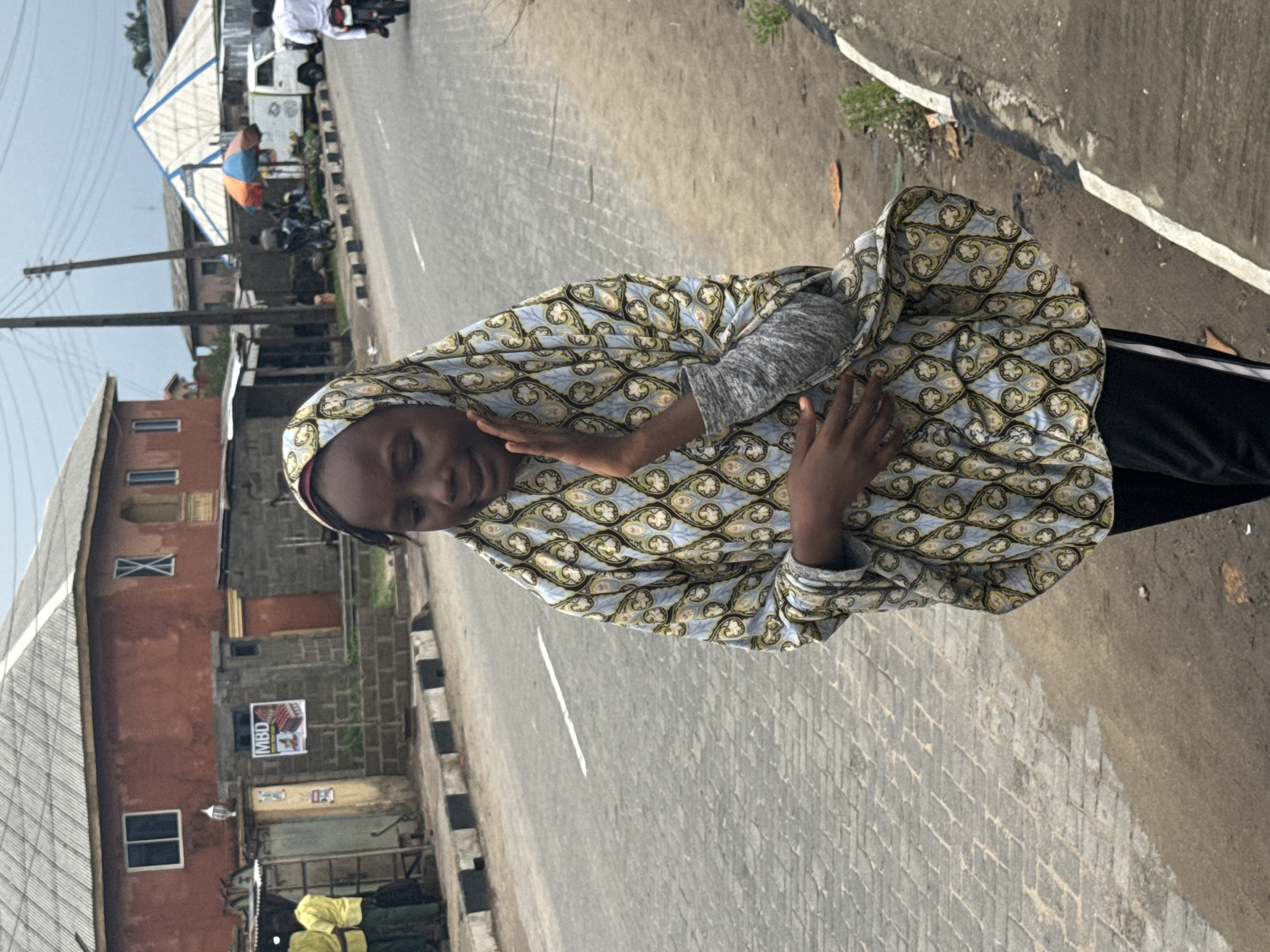 A woman wearing a patterned headscarf and robe stands on a paved street in Badagry, Nigeria. The street has a white line down the middle and black and white striped curbs. Behind the woman, there is a building with a sign posted on the side. In the background, there is a canopy, a white truck, and people on motorcycles. The sky is overcast.