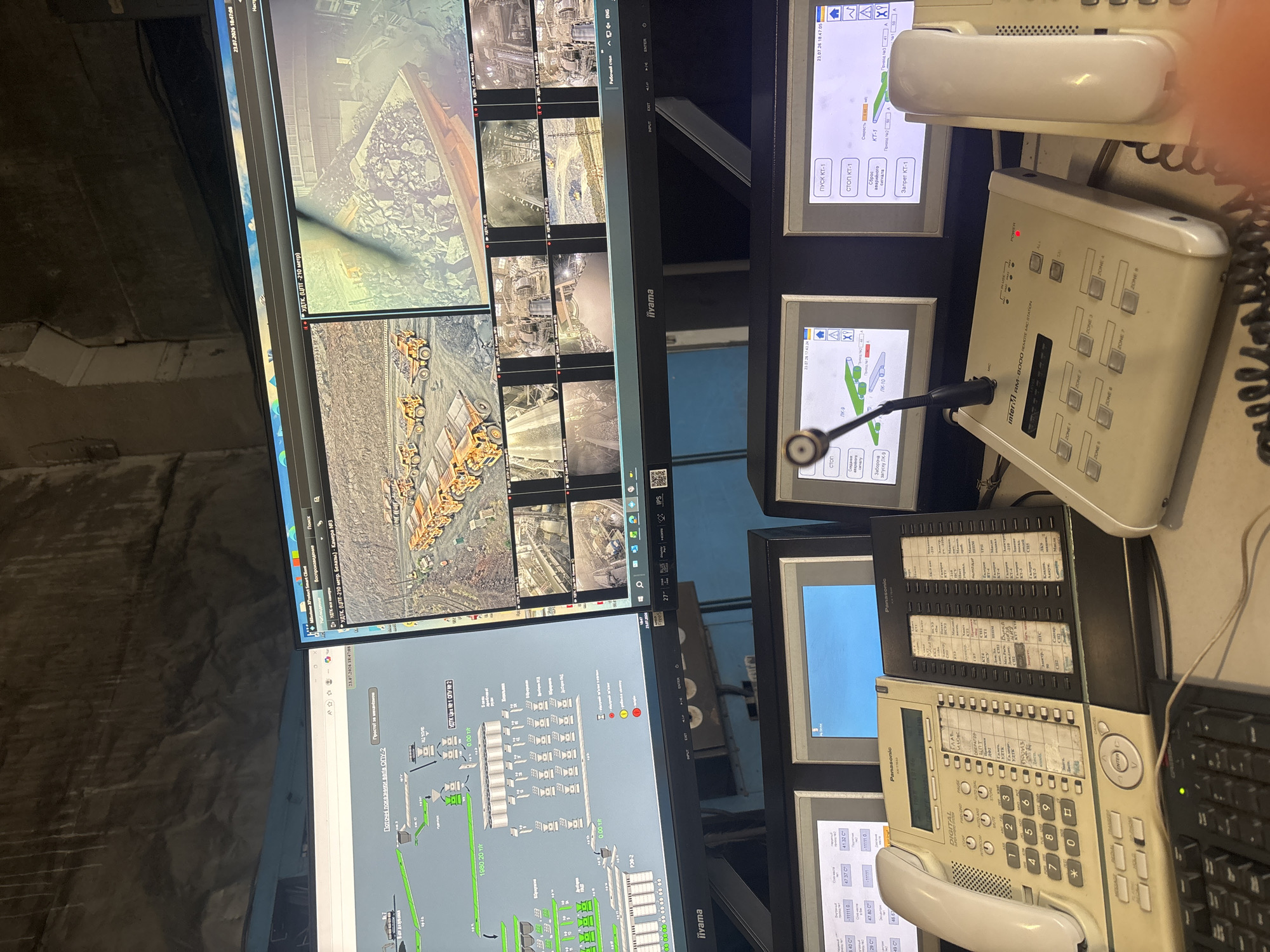 The image shows a control room with multiple monitors displaying what appears to be industrial processes and security camera feeds. The setting is indoors, likely a factory or mining operation, given the visual content on the screens. Several large monitors are arranged in a grid, showing schematics of conveyor belts, material processing equipment, and live video feeds from various locations, some of which feature heavy machinery like trucks and excavators.
On the right side of the image, there are two large display screens showing process statuses and controls, with Russian text indicating commands like "Start KT-1" and "Stop KT-1," and "Reset alarm signal." Below these screens are a Panasonic digital telephone and an intercom system, labeled "RM-8000 REMOTE MIC STATION" with various zone buttons. Another Panasonic phone is visible on top of a rack of equipment.
The visible text, primarily in Russian, suggests operational control and monitoring of an industrial facility. Dates and times are visible on some screens, such as "23.07.26 18:47:05" and "23.07.26 17:43:24," indicating a specific point in time. There are no people visible in the image, and the overall atmosphere is one of quiet, technical operation. The location context of Kryvyi Rih, Ukraine, suggests this control room is part of a mining or metallurgical complex in that region. The image captures a snapshot of a busy industrial control center.