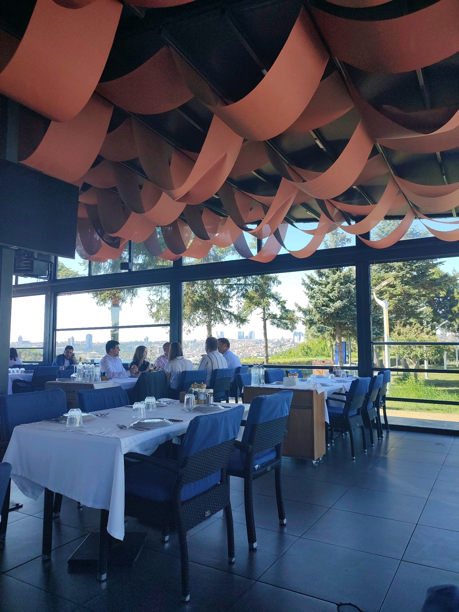 The image depicts the interior of a restaurant with large windows offering a view of Üsküdar, Turkey. The restaurant's ceiling is adorned with suspended, wavy orange fabric panels. Several tables are set for dining, covered with white tablecloths and blue chair cushions. In the background, through the windows, a city skyline is visible, with trees and greenery in the foreground. A group of people are seated at a table on the left side of the frame, engaged in conversation. The overall atmosphere suggests a contemporary dining establishment with a scenic backdrop.