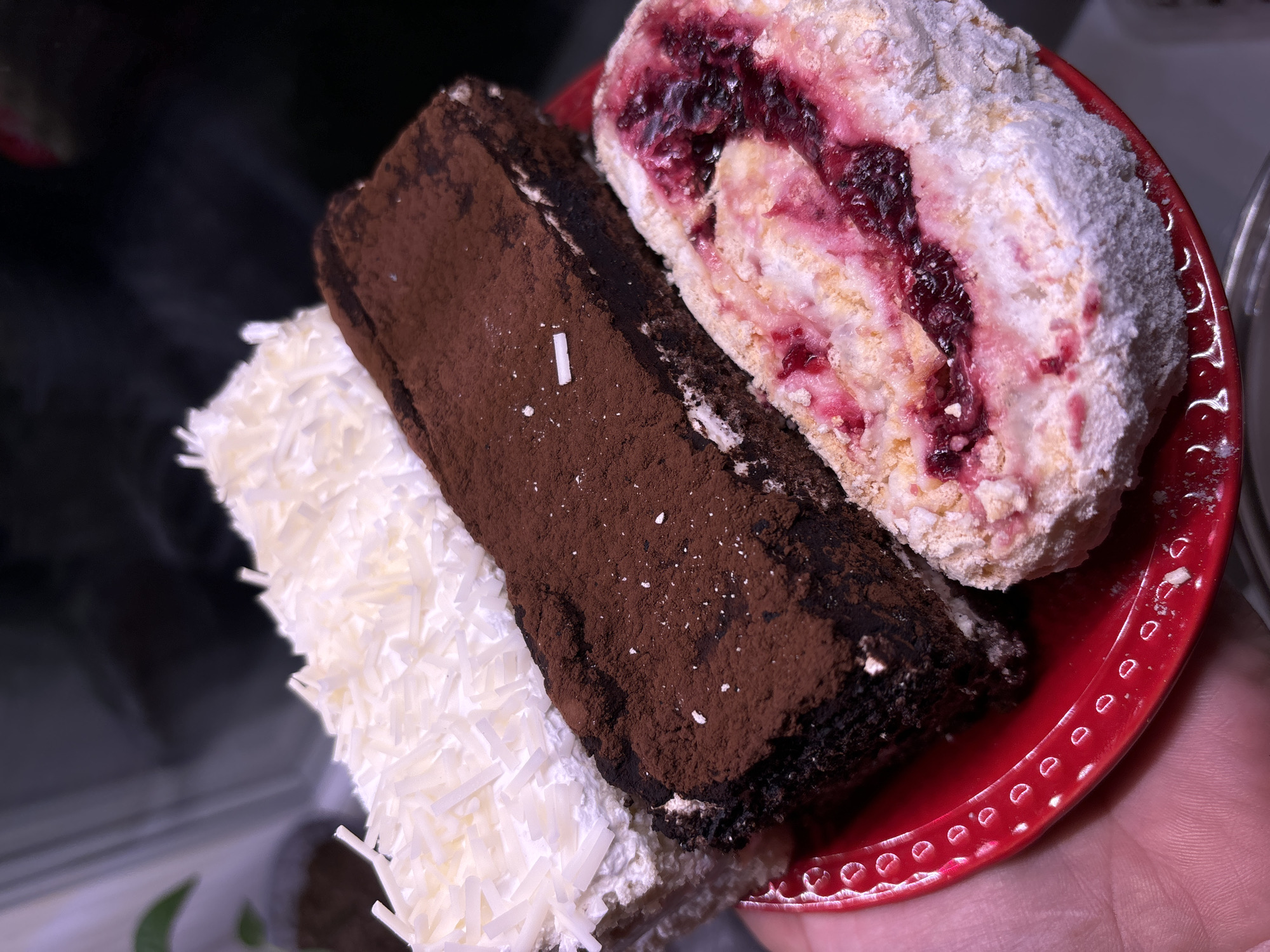 A close-up shot features three slices of different cakes arranged on a small red plate being held in a hand. From left to right, the slices are covered in white coconut shavings, cocoa powder, and powdered sugar, respectively. The cake covered in cocoa powder is rectangular, while the one with powdered sugar is round and has a red berry filling. The cakes are positioned close together, creating a stacked arrangement. The scene is brightly lit, highlighting the textures of the cakes. It is not possible to infer the city or any environmental details from the image.