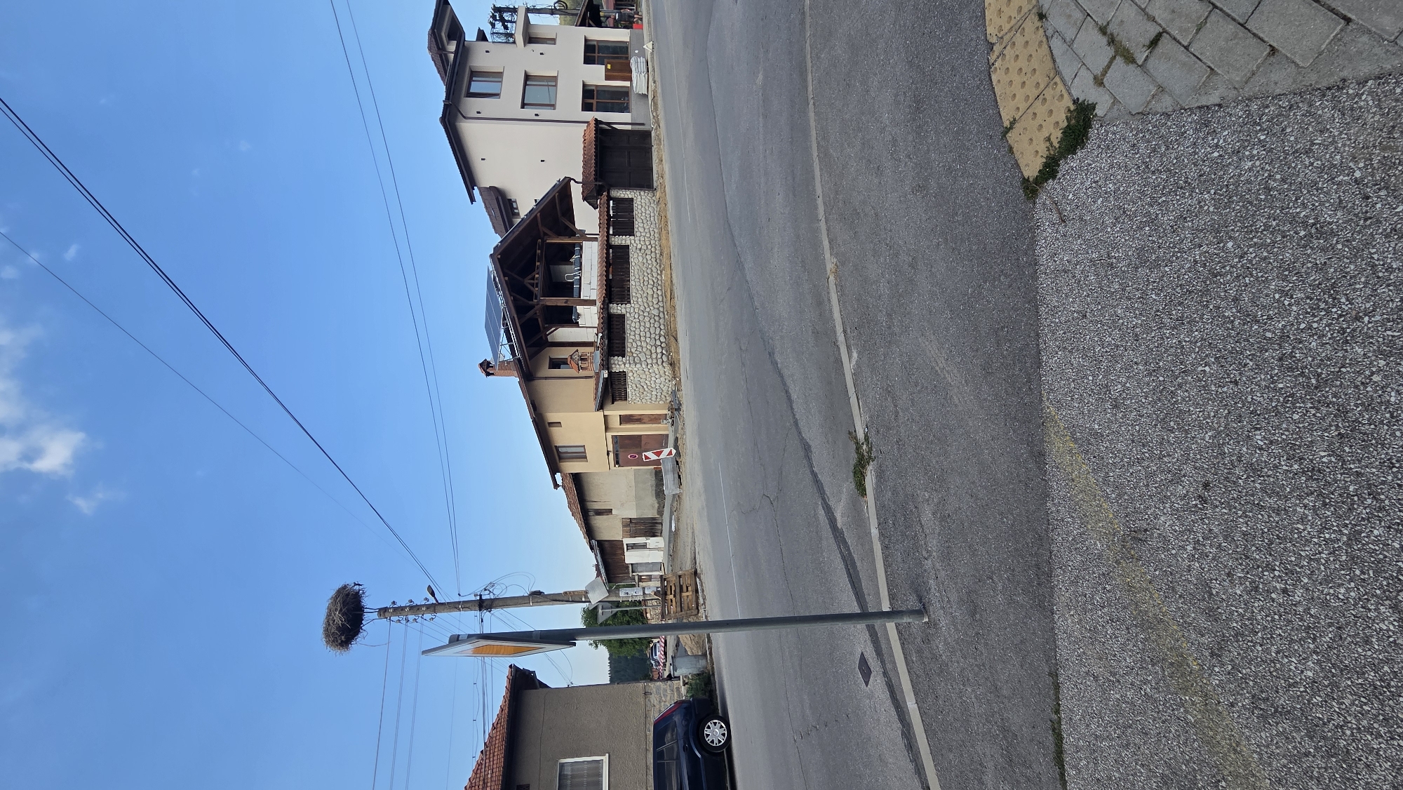 In the city of Bania, Bulgaria, a street scene unfolds under a clear blue sky with a few scattered clouds. On the left side of the image, a dark-colored SUV is parked near a two-story house with a red-tiled roof. A tall pole with a stork's nest sits prominently in the foreground, positioned between the houses and the road. The nest is a large, intricate structure of twigs and other materials.
Across the asphalt road, a series of buildings are visible. The first is a single-story, light-yellow house with a chimney and solar panels on its roof. This house is partially enclosed by a low stone wall. To its right, a taller, modern beige building with multiple windows and balconies stands. Construction materials, including stacks of concrete blocks, are present near the beige building.
The road itself is paved with asphalt, showing some cracks and patches. The foreground of the image features a grey, granular surface, transitioning into a sidewalk with interlocking pavers and tactile paving blocks. The overall impression is that of a residential area on a bright day.