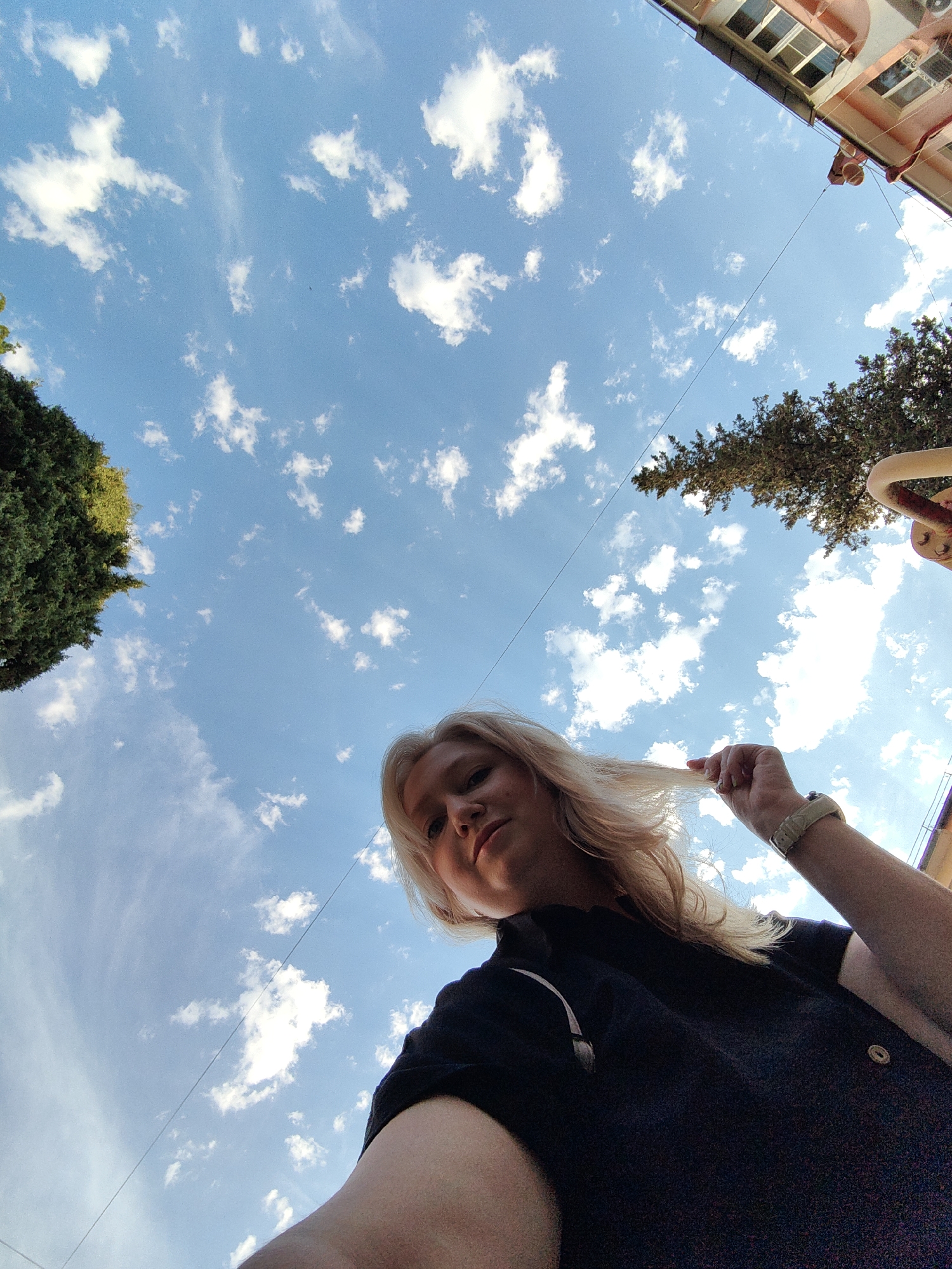 A low-angle, full-body shot captures a young woman with blonde hair playing with her hair against a bright blue sky filled with fluffy white clouds. She is wearing a black shirt and a light-colored watch. To the left of the frame, a tall, green tree is visible. In the upper right corner, a building with pink walls and multiple windows can be seen. The image appears to have been taken outdoors, possibly in a city park or street in Sochi, Russia.