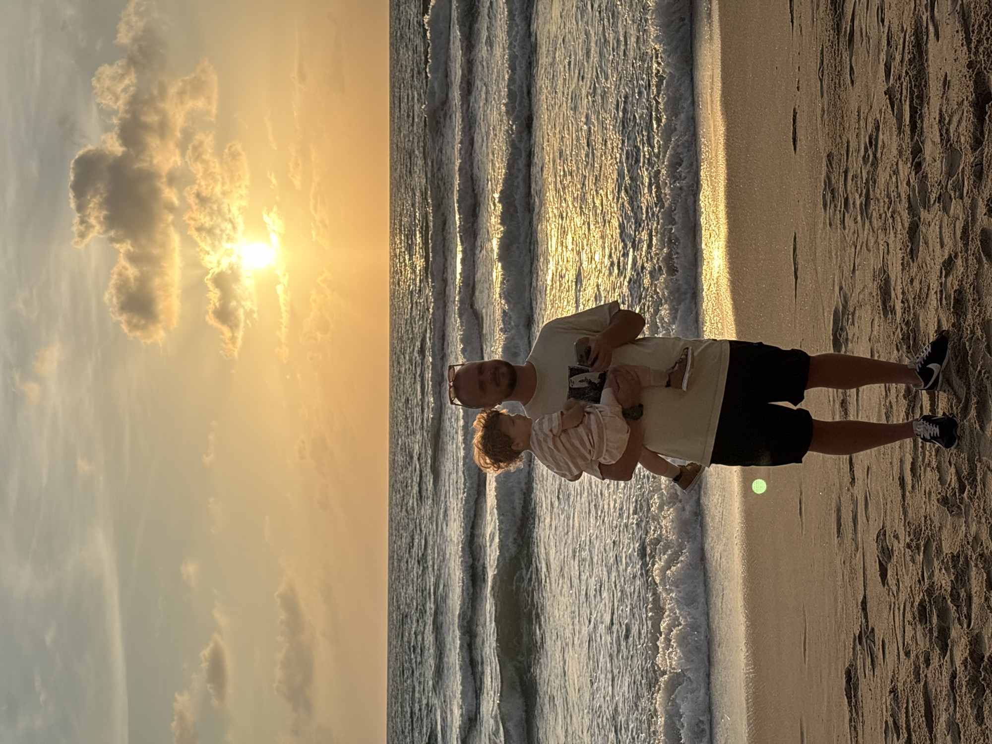 A man stands on a sandy beach in Choeng Thale, Thailand, holding a young child. The man, wearing a t-shirt and dark shorts, has sunglasses perched on his head and is holding a mobile phone. The child, dressed in a striped outfit, is being held in his arms. They are facing the ocean, with waves gently rolling onto the shore. The sun is setting, casting a warm orange glow across the sky and reflecting on the water. Clouds are visible in the sky, illuminated by the setting sun. The sand on the beach has footprints and is illuminated by the sunset.