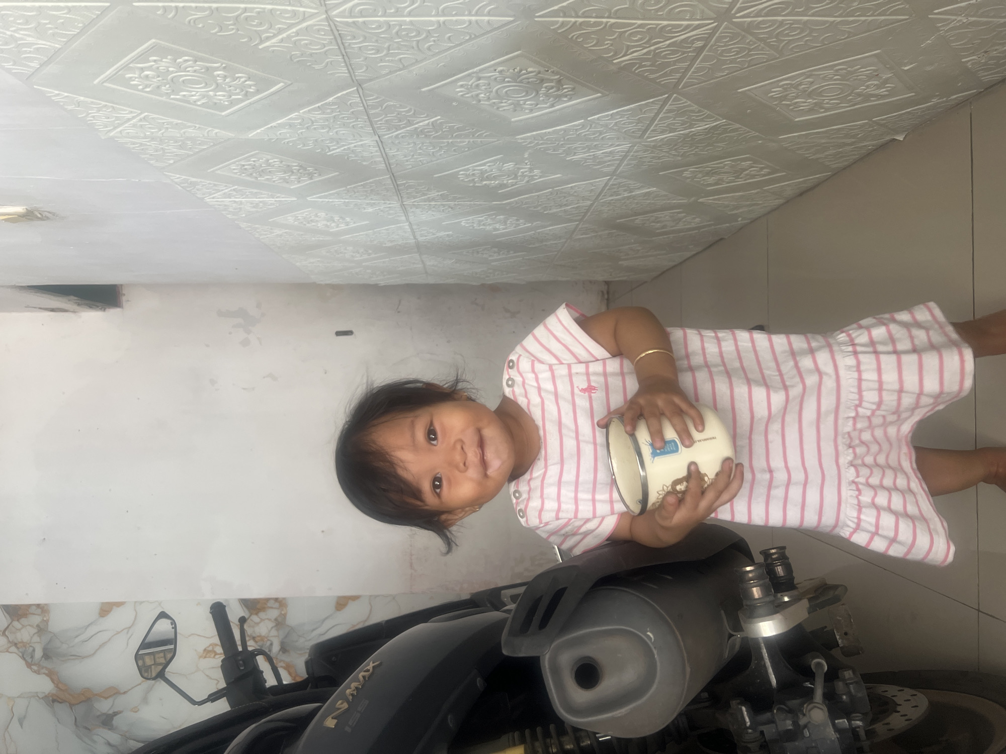 A young Southeast Asian child, likely a girl due to her attire and accessories, stands holding a white mug with pink stripes and a decal of a cartoon character. She is wearing a pink and white horizontally striped dress with ruffled hem and shoulder buttons. Her left wrist bears a thin gold bracelet. The child has dark hair and is smiling, with some white residue on her upper lip. She is positioned in what appears to be an outdoor or semi-enclosed area, with a light-colored tiled floor. To her left, a dark-colored motorcycle is partially visible, featuring a rear wheel with a brake disc and part of its exhaust system. The background includes a wall with a marble-patterned section on the left and a white wall with a raised diamond-shaped textured pattern on the right. The lighting suggests it is daytime. The scene is likely in an urban or suburban residential area.