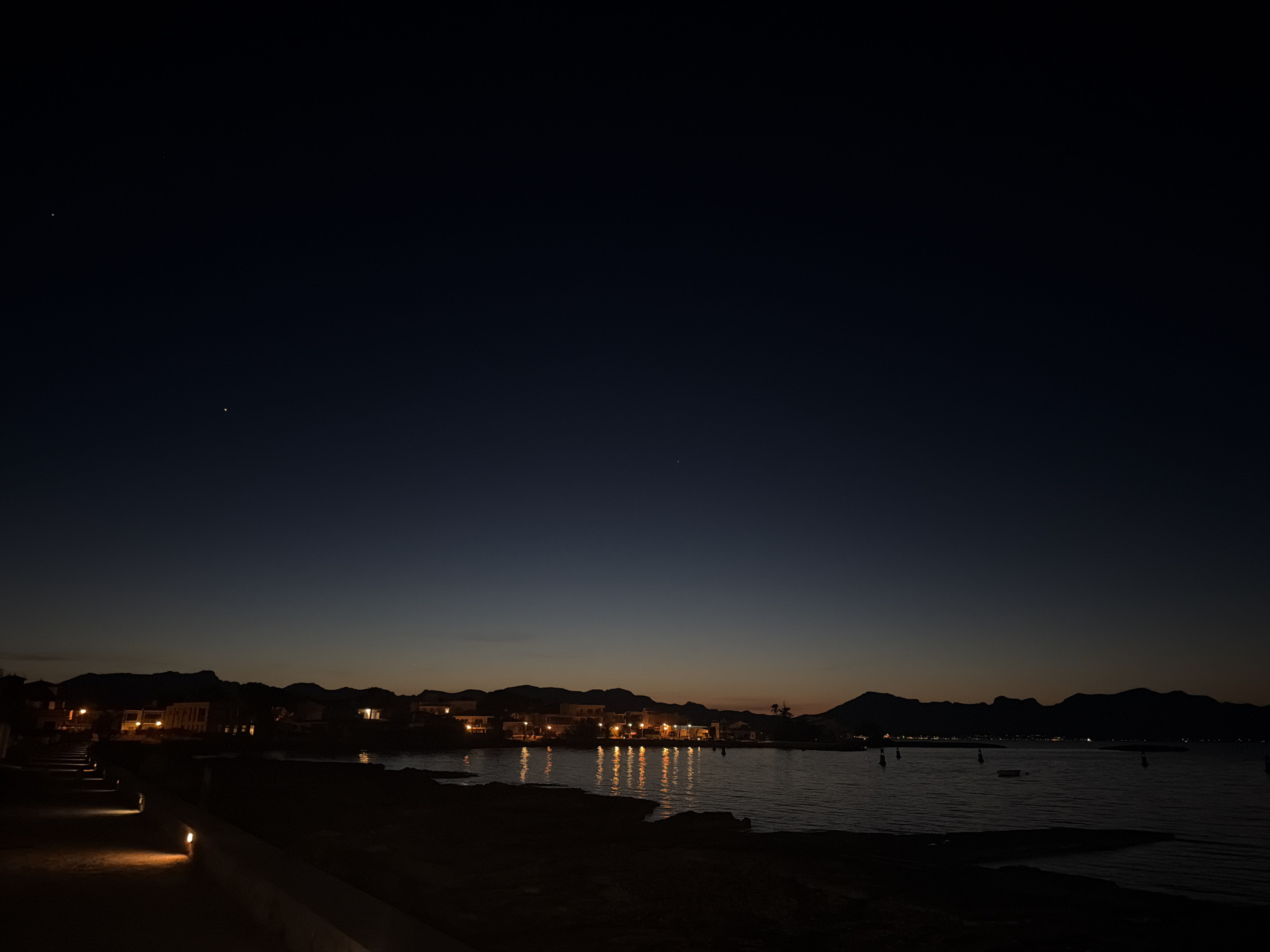 At dusk in Alcúdia, Spain, a body of water reflects city lights. In the sky, two small points of light appear to be stars. Houses are visible along the distant shoreline. The nearest ground area is dark, with a lit walkway on the left.