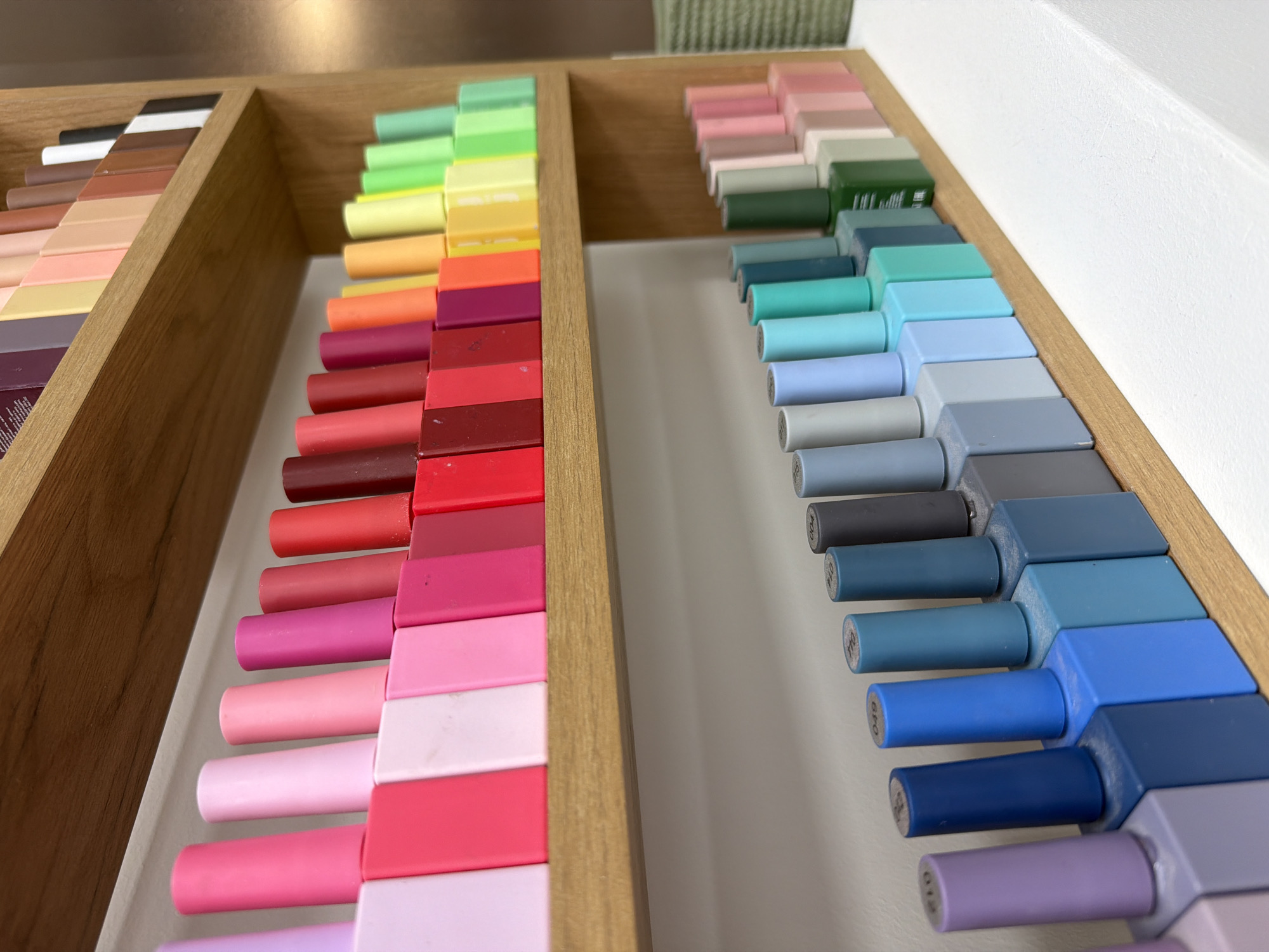 The image shows a close-up view of two wooden trays containing rows of nail polish bottles arranged in color gradients. The tray on the left displays nail polishes in shades of brown, pink, red, orange, yellow, and green. The tray on the right features nail polishes in shades of pink, green, teal, blue, gray, and purple. The bottle caps are square and uniform in size, while the bottles themselves have cylindrical stems. The image is taken from a high angle, showcasing the variety of colors and arrangement of the nail polishes. There is no evidence to suggest this image was taken in Moscow, Russia.