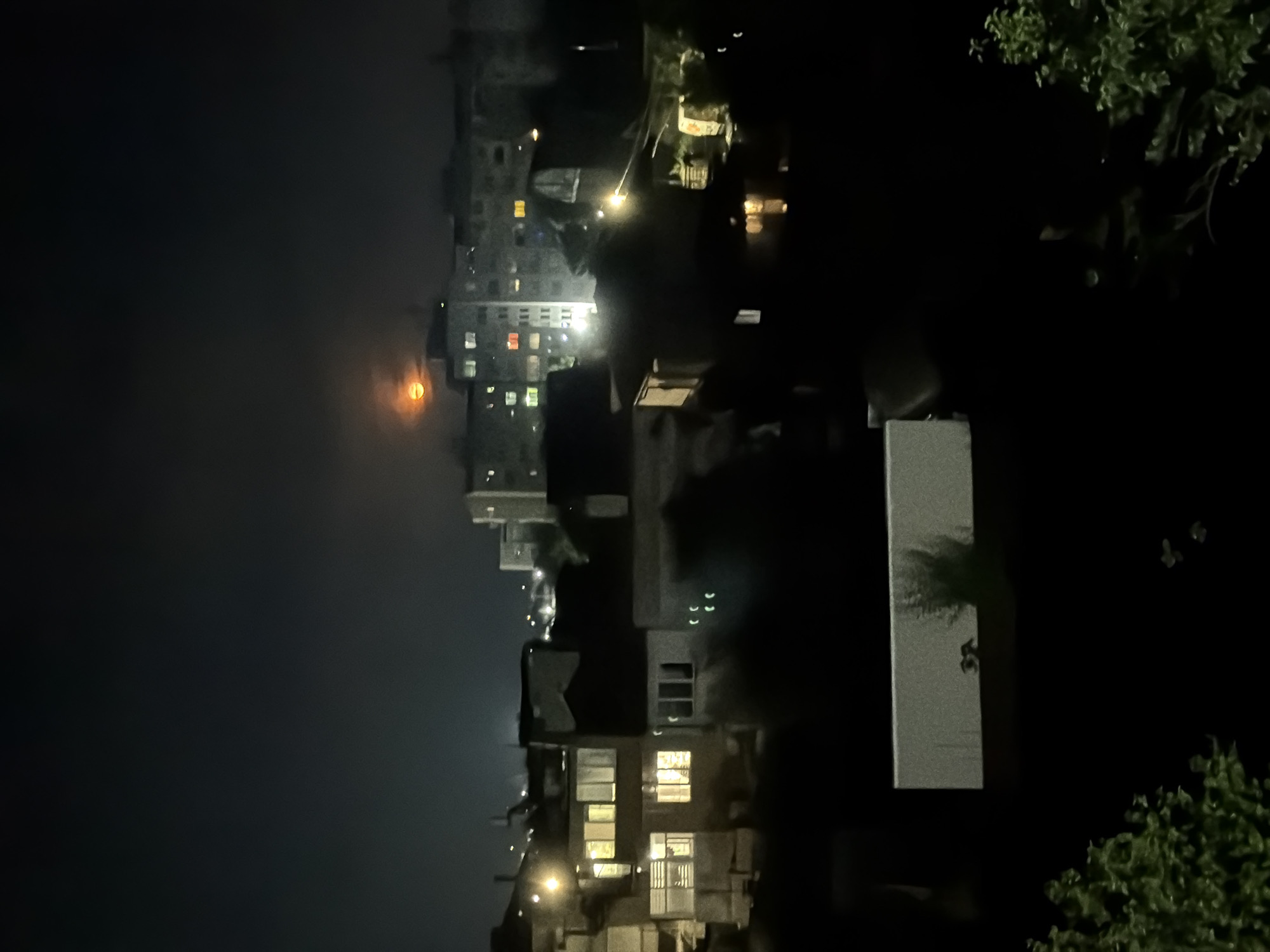 The image captures a nocturnal urban landscape in Burgas, Bulgaria, dominated by a large, reddish-orange moon partially obscured by atmospheric haze or light clouds on the left side of the frame. The scene is illuminated by the moon and numerous artificial lights from buildings.
In the mid-ground and extending to the right, a cluster of residential or mixed-use buildings stands. Many windows in these multi-story structures are lit with warm, yellow, and sometimes cooler white lights, suggesting active occupancy. Several buildings have distinct architectural features, including visible balconies and varying rooflines. Streetlights or building exterior lights also cast bright glows, creating points of intense illumination within the dark city fabric.
On the far right and in the lower right corner, the dark silhouettes of trees with visible green foliage are present, adding a natural element to the urban setting. The sky is dark, indicating a clear night, but the atmospheric conditions around the moon suggest either haze, light cloud cover, or particulate matter in the air contributing to its striking reddish hue. No specific human activity or identifiable individuals are visible, presenting a quiet, moonlit cityscape. No legible text is visible in the image.
