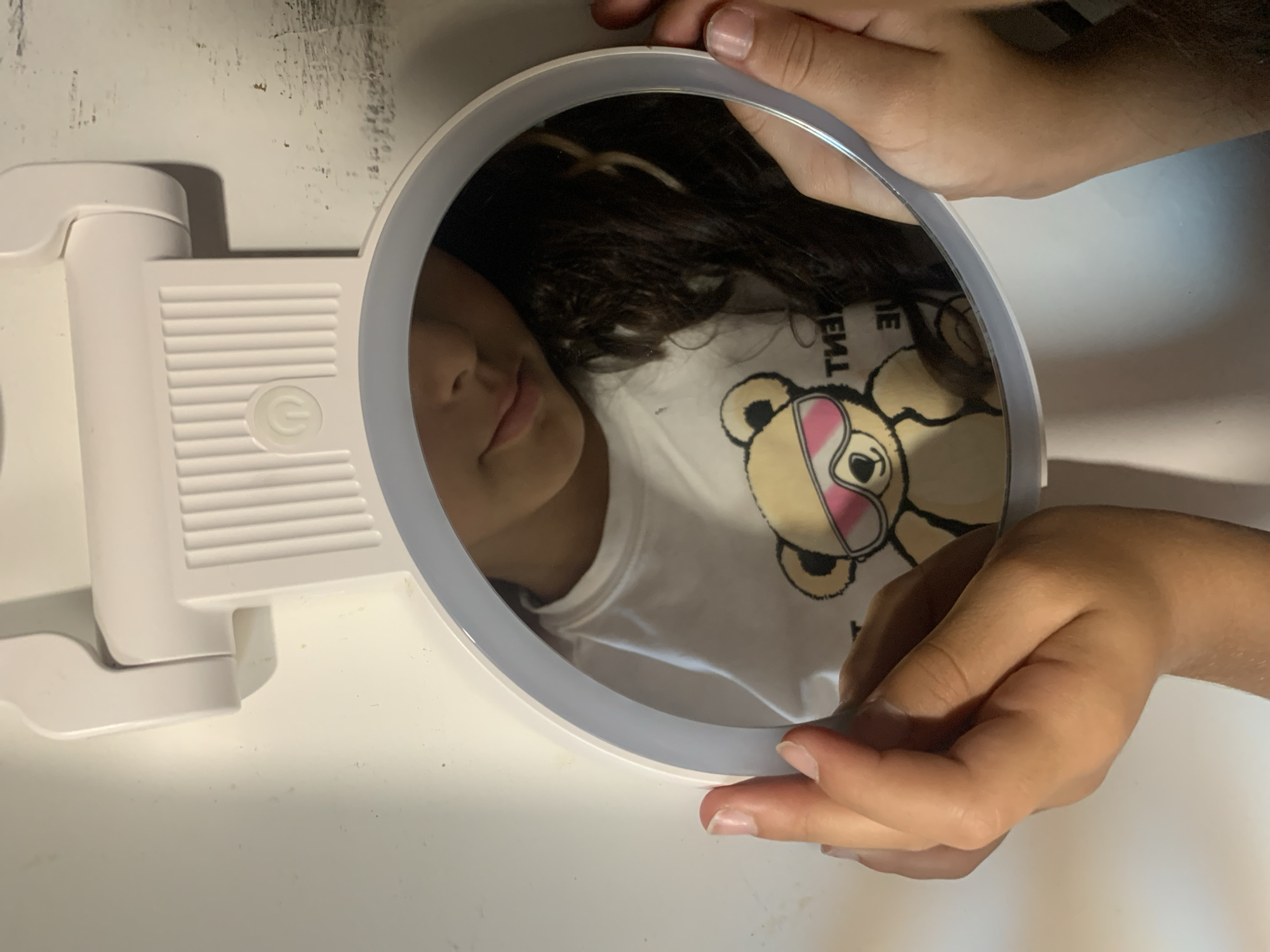 A close-up overhead shot shows a young person's face reflected in a circular mirror. The person is wearing a white t-shirt with a teddy bear graphic that has sunglasses. The mirror is held by the person's hands, one on each side. The background appears to be a white surface with some texture and discoloration. The lighting suggests an indoor environment, possibly a desk or table. No specific location in San Quirico d'Orcia, Italy is discernible from the image.
