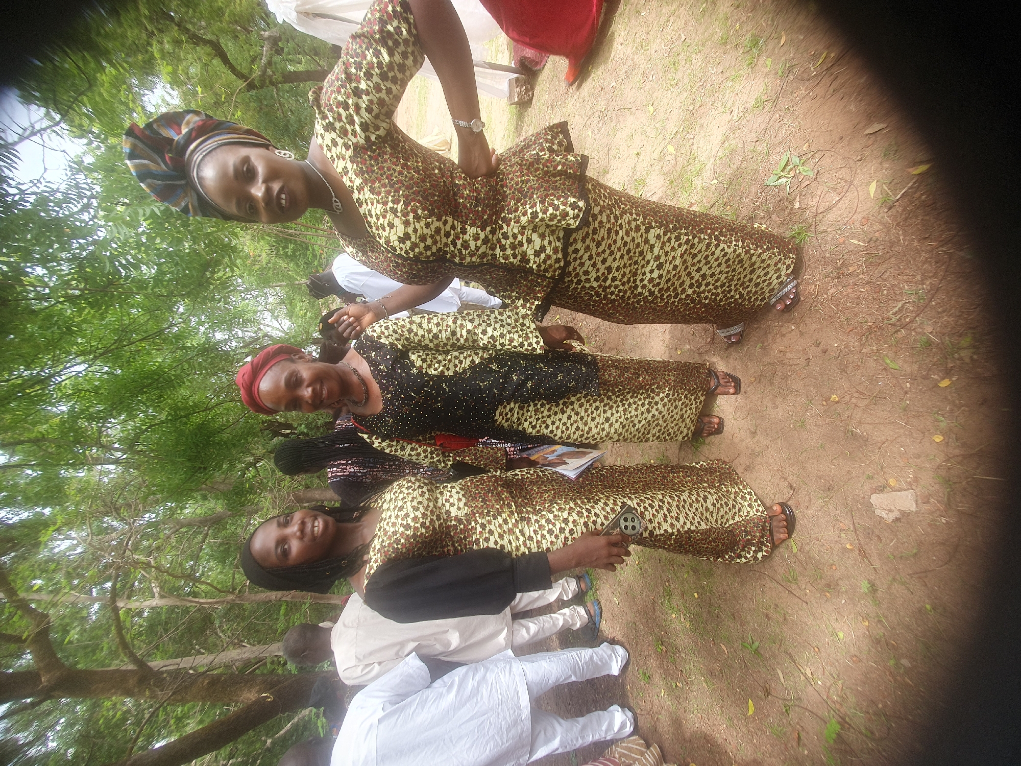 Three women stand in a grassy area in Unknown, Unknown. The women are wearing similar dresses patterned with brown and yellow designs. The first woman, located at the top of the image, has a patterned headwrap, and a watch on her left wrist. The middle woman has a red headwrap and a black section on her dress. The third woman, located at the bottom of the image, holds a cell phone in her right hand. Several people in white clothing are standing near her. Trees and foliage are visible in the background. The ground is dirt and grass.