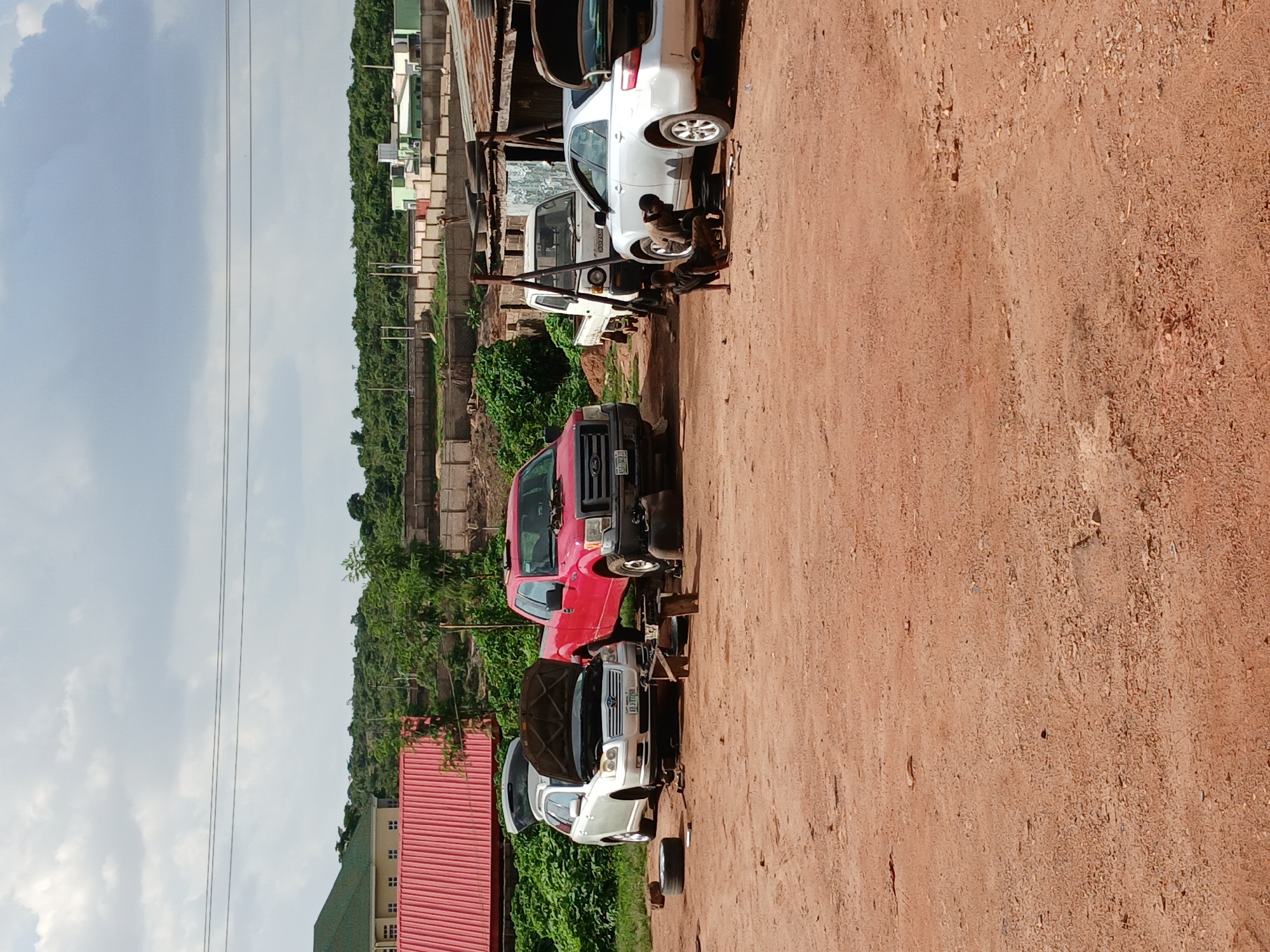 The image shows an outdoor scene with several vehicles being worked on. In the foreground, a red pickup truck is parked. Behind it, a silver sedan with its hood open is on a jack, with tools scattered around it. Two individuals, appearing to be mechanics, are actively working on the car. Further back, a white van and another white vehicle are visible. The scene appears to be a car repair or scrapyard. The ground is covered in red dirt, suggesting a dry environment. In the background, there are buildings and lush green vegetation, with a hilly landscape under a cloudy sky. The lighting suggests it is daytime. A red corrugated metal building is partially visible on the left. The overall atmosphere seems to be one of active work and maintenance. A license plate on the silver car reads "ARJ 7709".