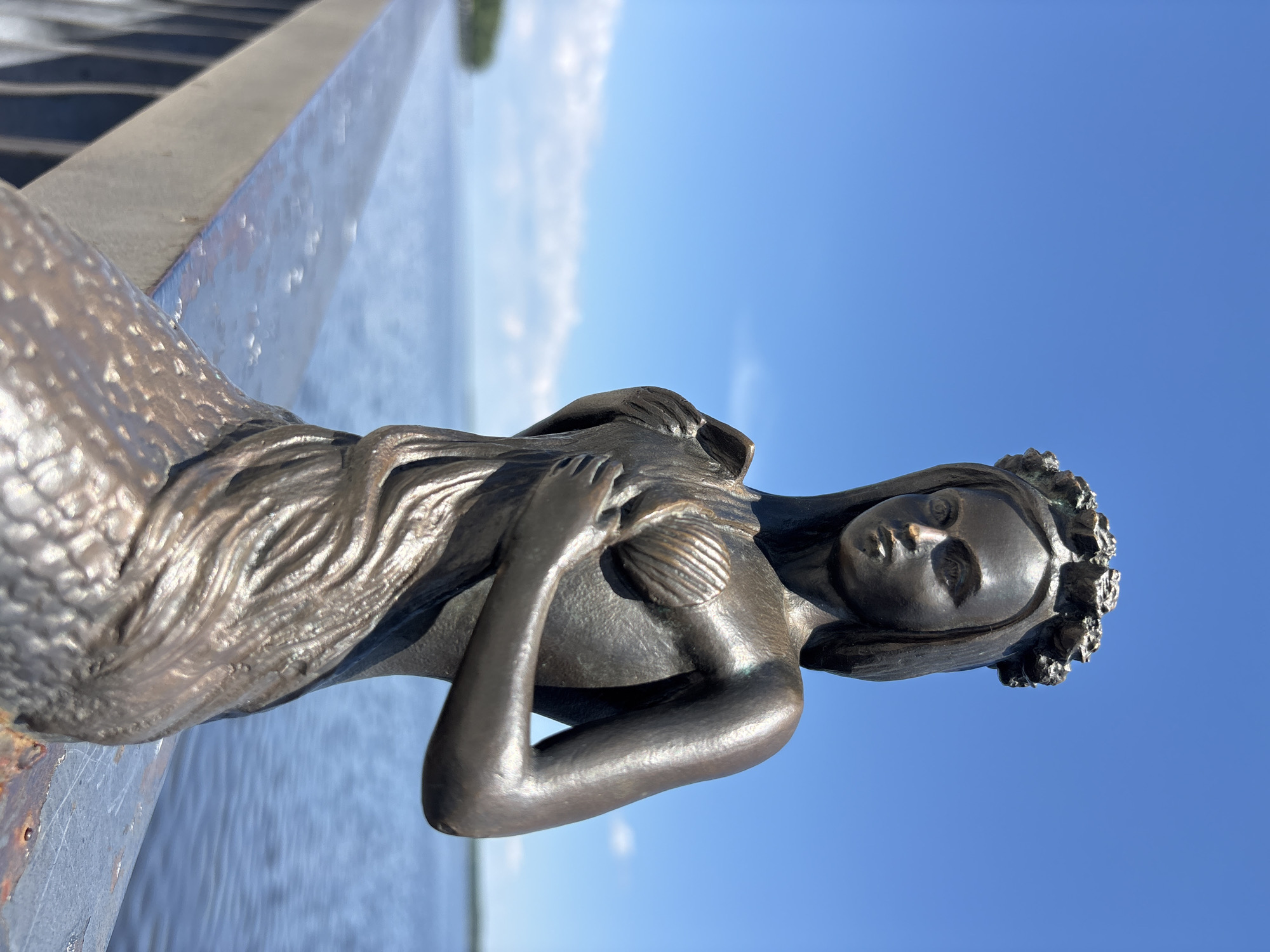 A bronze statue of a mermaid sits on a metal railing overlooking a body of water under a clear blue sky. The mermaid has a crown of flowers on her head and holds a shell to her chest. Her lower body is scaled like a fish. The water is calm and reflects the sky. In the distance, there is a faint line of trees on the horizon. The location is the embankment in Chaikovsky, Russia, a city situated on the Kama River.