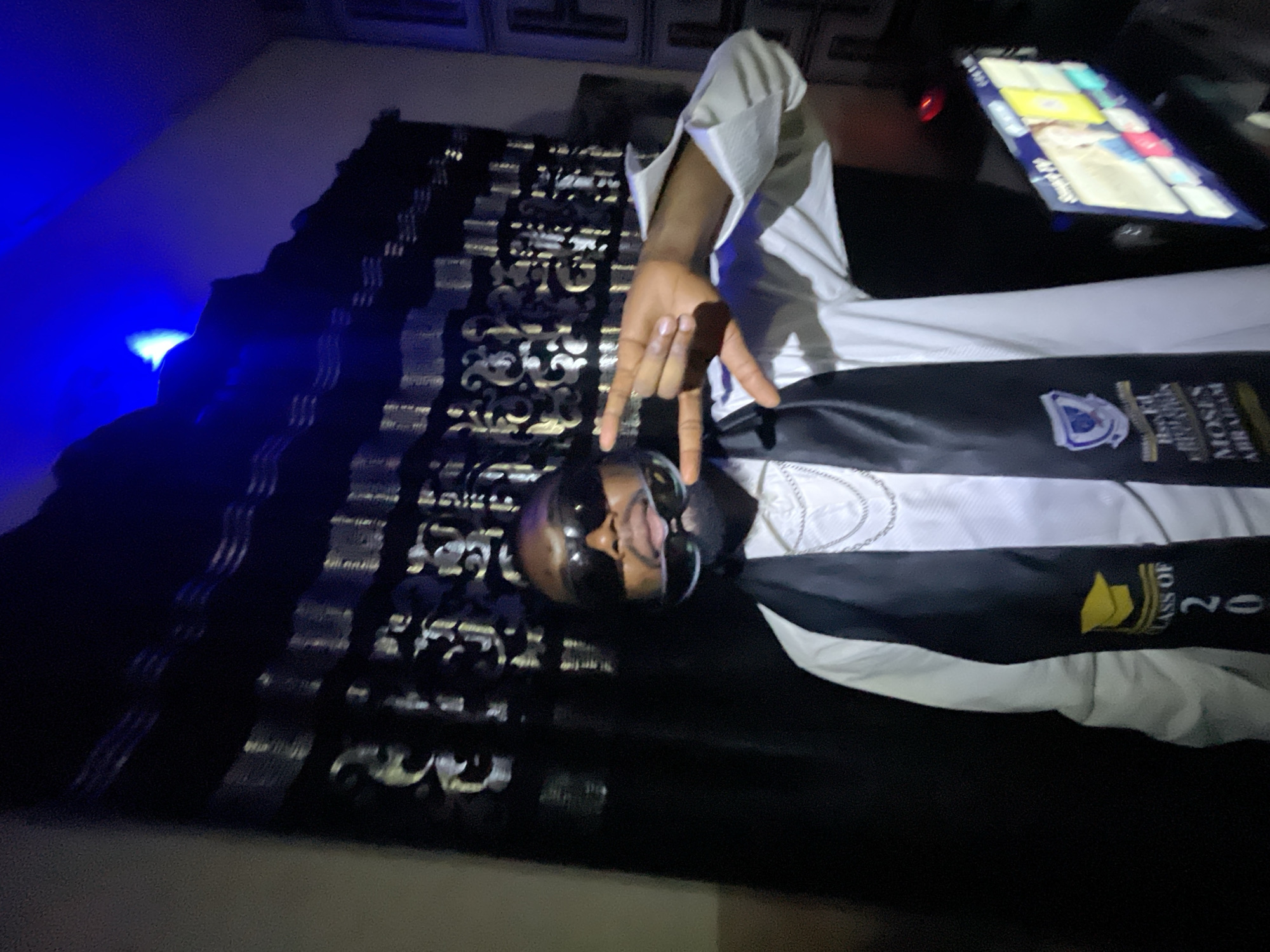 A person with a beard and sunglasses is lying on a bed with black and white patterned sheets, wearing a white shirt and a black graduation stole with "CLASS OF 20" printed on it. The stole also features a school emblem and text appearing to include the name "MOSES ABRAHAM". The person is making a hand gesture resembling "hook 'em horns". There's a blue light in the background and a device with a screen showing a user interface is visible on the upper right. The location is not evident from the image.
