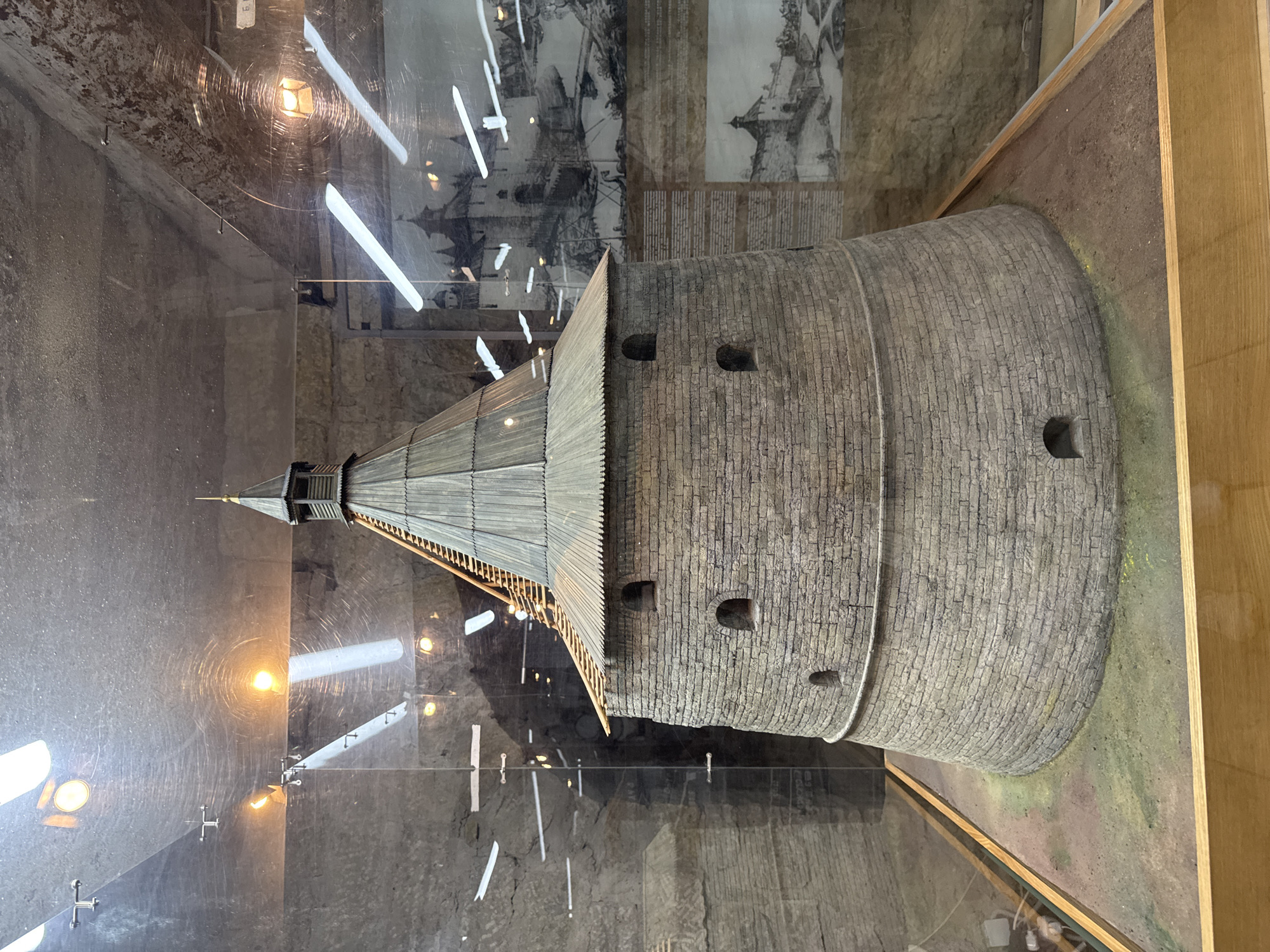 This media file shows a detailed diorama of a cylindrical stone tower with a wooden roof. The tower is constructed from rough-hewn bricks and has several small, dark openings that appear to be windows or embrasures. The wooden roof is pointed and has a layered, shingled appearance, topped with a slender spire. A narrow wooden walkway runs around the base of the wooden roof structure.
The diorama is housed within a glass enclosure, which reflects the surrounding environment. The reflections reveal overhead lighting fixtures and the interior of what appears to be a museum or exhibition space. In the background, behind the diorama, is a large, black and white illustration or mural depicting a historical scene, possibly a battle or siege, with ships and fortifications. There are also some unintelligible lines of text visible in the background.
The diorama is set on a base that simulates ground, covered with brown soil and patches of green and yellow moss or vegetation. The overall impression is that of a historical model, likely representing a fortress or defensive tower, located in Shlisselburg, Russia, as suggested by the location context. The lighting suggests an indoor setting, likely during daytime or with artificial lighting. There are no people visible in the media file, and no specific emotions are conveyed.