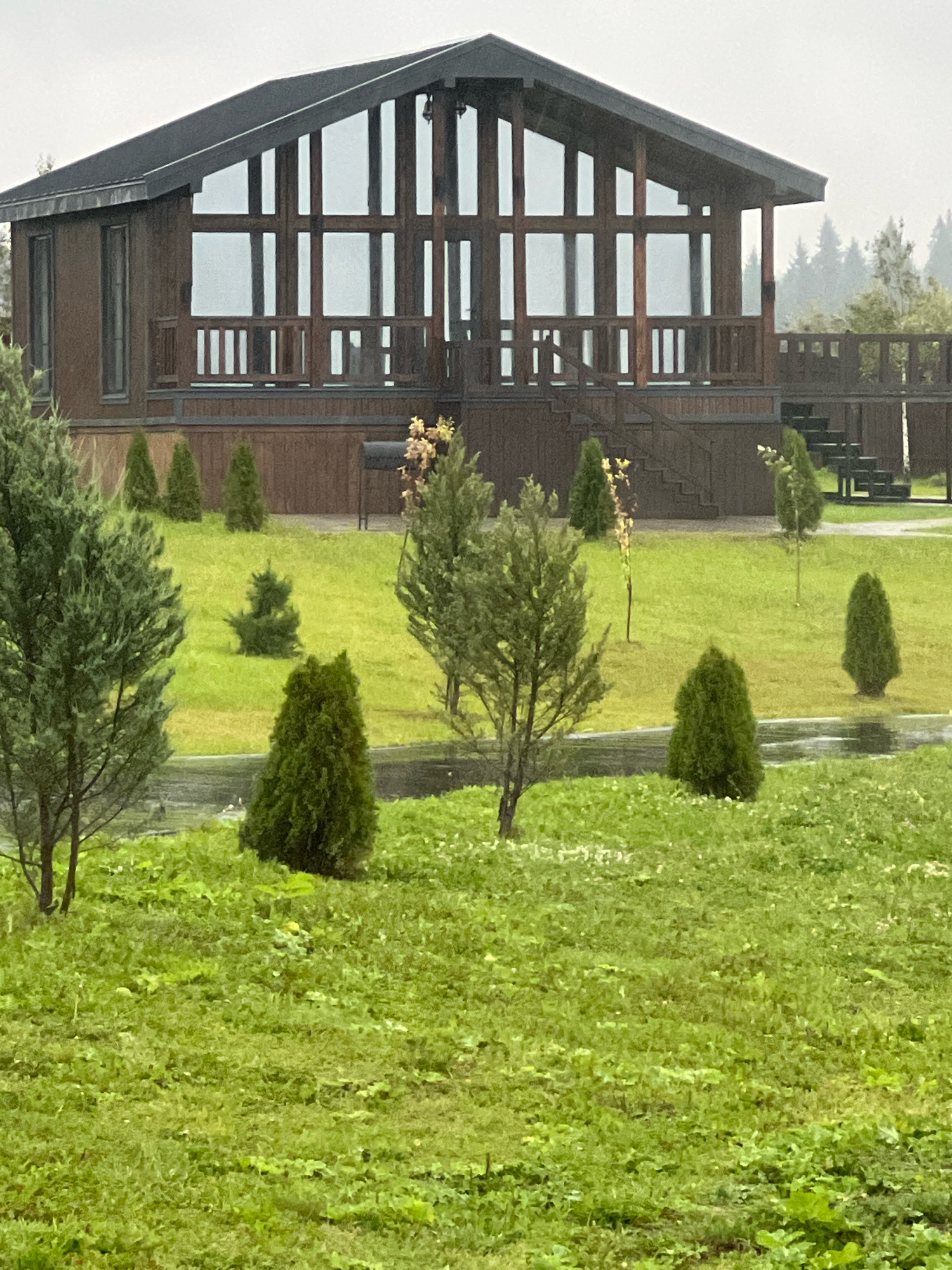 The image shows a modern wooden house with a large glass facade, located in a rural or suburban area with a lawn and trees. The house has a dark-colored roof and wooden siding. There are stairs leading up to a deck or porch that wraps around part of the house. Several evergreen trees of varying sizes are scattered across the lawn. A small grill is visible on the deck of the house. The ground appears wet, suggesting recent rain. The background shows more trees and a hazy sky. There are no people visible in the image.
