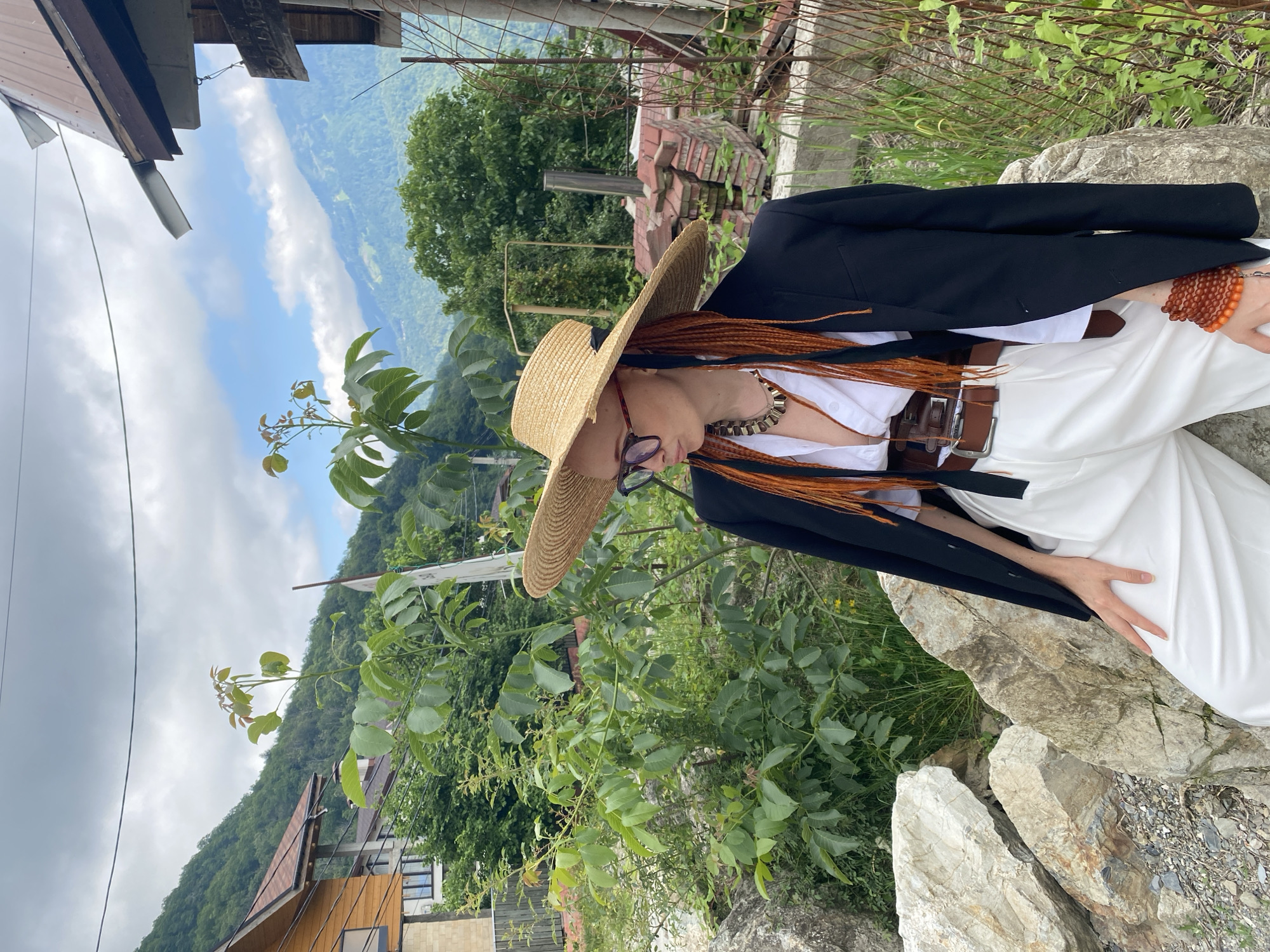 A person with long, braided auburn hair and a wide-brimmed straw hat is seated on a rocky outcrop, looking down. They are wearing a white shirt, white pants, a black blazer, and a brown belt. A thick, beaded necklace and a bracelet made of amber-colored beads are visible. The person's left hand rests on their lap.
The background depicts a lush, green mountainous landscape under a cloudy sky. In the distance, rolling hills covered in trees can be seen, with a valley in between. Some houses with pitched roofs are nestled on the lower slopes. In the foreground, to the left, is a section of wooden siding and a metal pipe, suggesting a structure nearby. Green foliage and rocks dominate the immediate surroundings. The lighting indicates it is daytime, likely overcast due to the clouds. The setting appears to be a rural or mountainous area, consistent with the location context of Krasnaya Polyana, Russia. There is no visible text in the image.