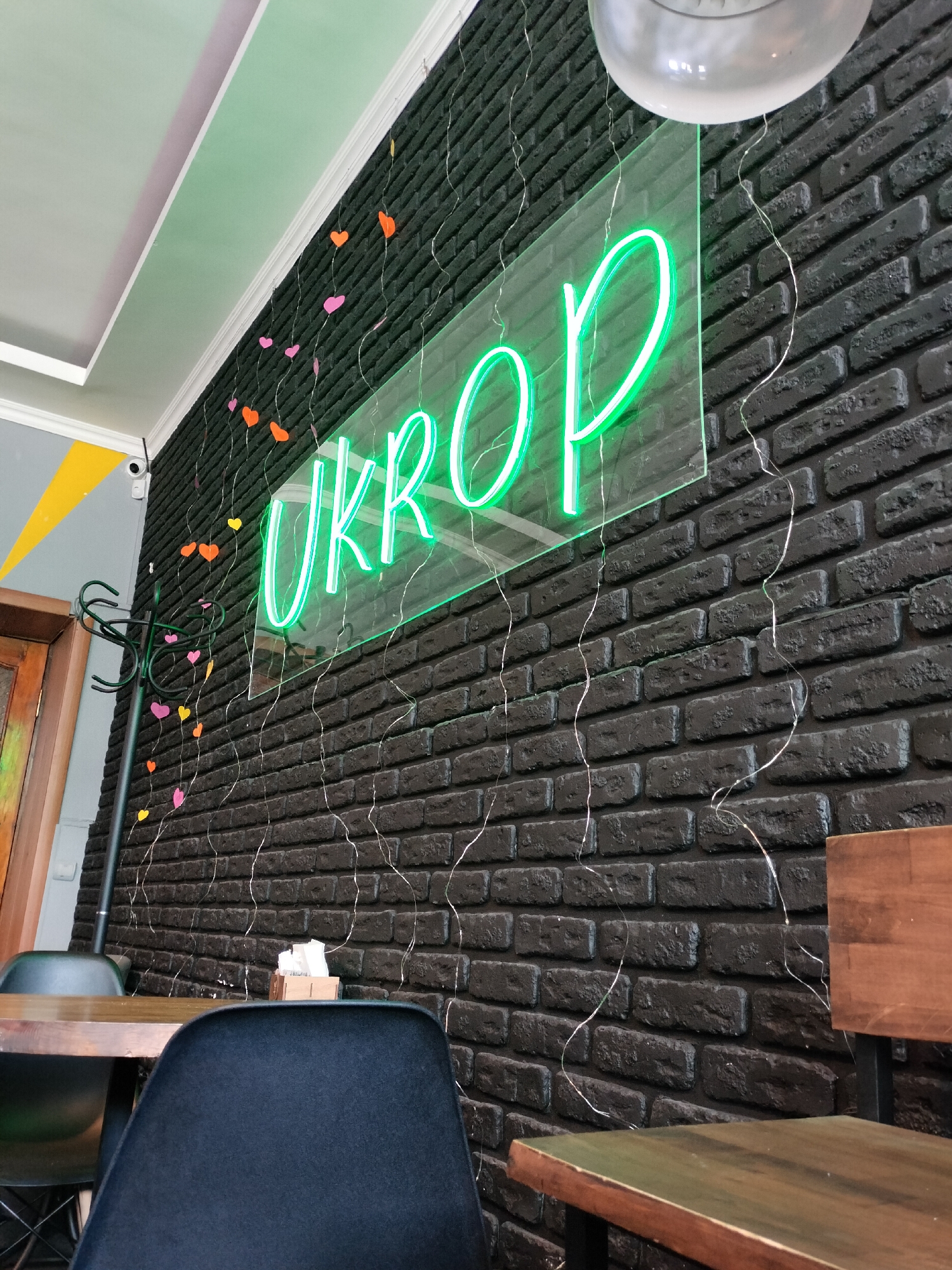The interior of a restaurant or café is depicted in the image. A black brick wall serves as a backdrop, adorned with a neon green sign that reads "UKROP." Strings of fairy lights with small, colorful paper hearts are draped along the wall. To the left, a coat rack stands near a partially visible doorway. A table and chair are in the foreground, suggesting a seating area for patrons. The overall ambiance seems casual and inviting.