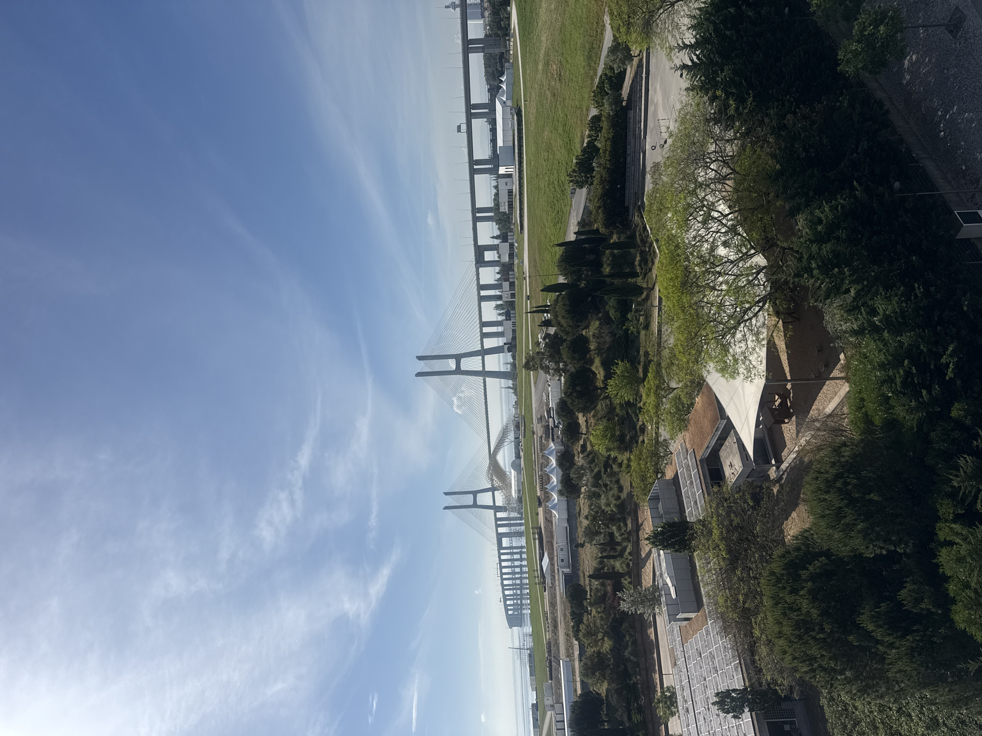 The image presents a panoramic view dominated by a vast suspension bridge stretching across a wide expanse of water under a clear blue sky. The scene is brightly lit by daylight, indicating mid-morning to early afternoon, with scattered wispy white clouds high above.
The impressive bridge, likely the Vasco da Gama Bridge in Loures, Portugal, features multiple towering pylons and a complex web of support cables. It extends horizontally across the frame, connecting the visible foreground land with a faintly discernible distant shore, where outlines of buildings or an industrial area can be seen. Below the bridge, the calm body of water reflects the bright sky.
In the foreground, to the right of the bridge, lies a sprawling green landscape. This area consists of well-maintained grassy lawns and a diverse collection of trees, ranging from light-foliaged deciduous trees to dense evergreen conifers. Interspersed within this park-like setting are several contemporary low-rise buildings and structures. Some buildings have distinctive white, tent-like roofs, while others appear more functional, with sections that resemble solar panel installations. A paved area, possibly an outdoor sports court with basketball hoops, is also visible among the greenery. Paved pathways and roads crisscross the landscape.
No individuals or explicit activities are visible, emphasizing the grandeur of the landscape and architectural elements. There is no discernible text in the image.