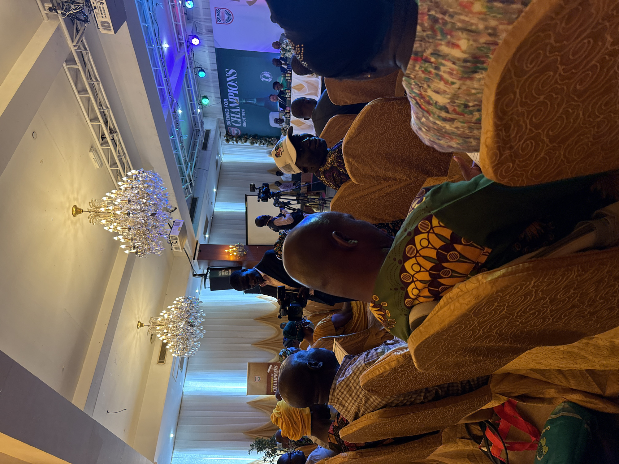 The image shows a room in Itirin, Nigeria set up for a formal event. Attendees are seated in upholstered chairs. At the front of the room is a stage with a banner reading "Brewed for Champions since 1974". There are cameras and personnel operating them, suggesting a recording or broadcast. Two large crystal chandeliers illuminate the room. The ceiling has exposed structural supports with stage lighting.
