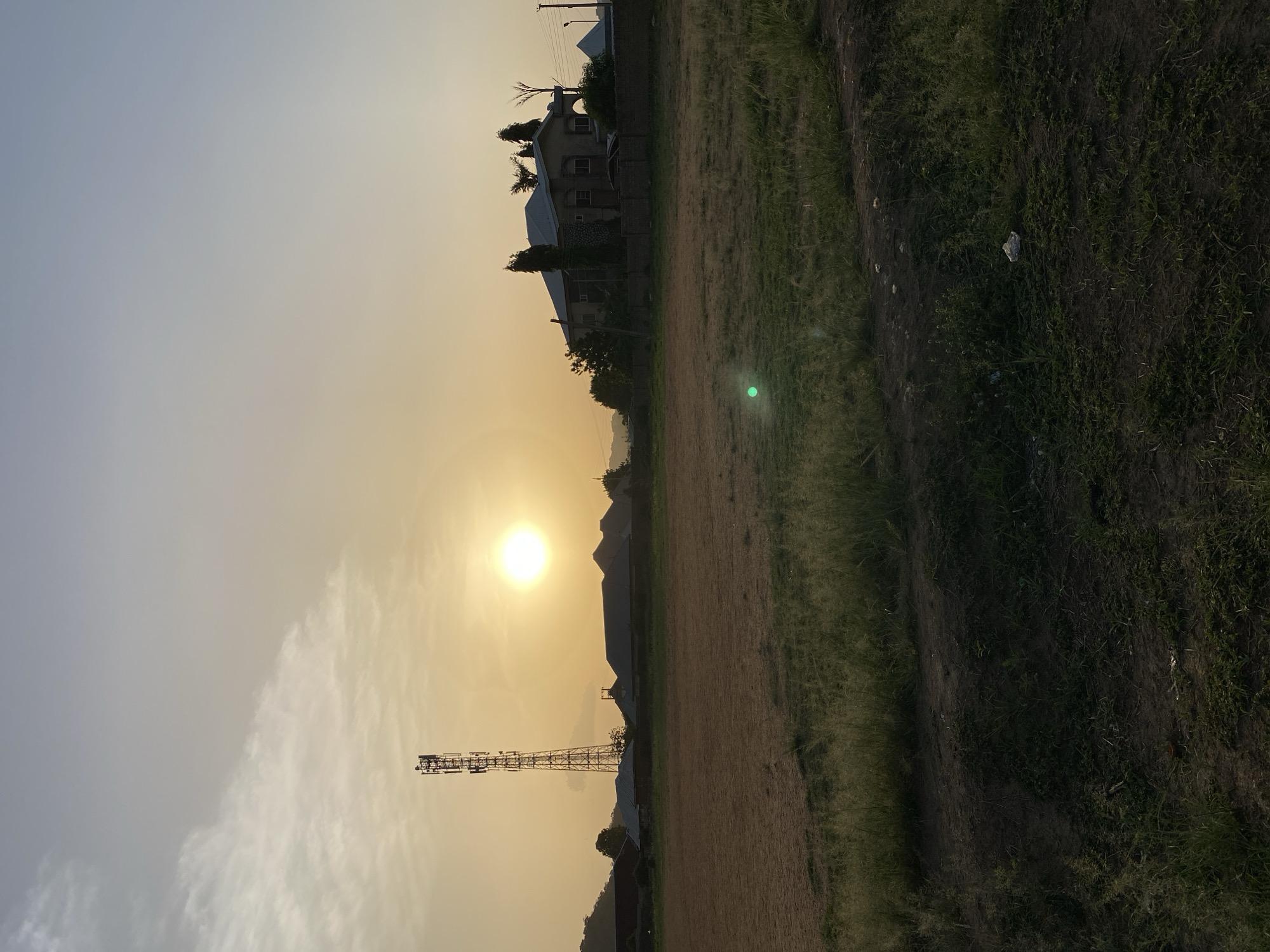 The image shows a landscape scene in Bukuru, Nigeria, featuring a grassy field with a dirt path running along the right edge. A communication tower stands prominently in the middle-left foreground, silhouetted against a bright, hazy sunset. The sky is a gradient of light gray to yellow. In the middle ground, there are houses and vegetation. The overall tone is warm and muted due to the lighting conditions. A glint or reflection is visible on the field. The image is rotated 90 degrees counter-clockwise.