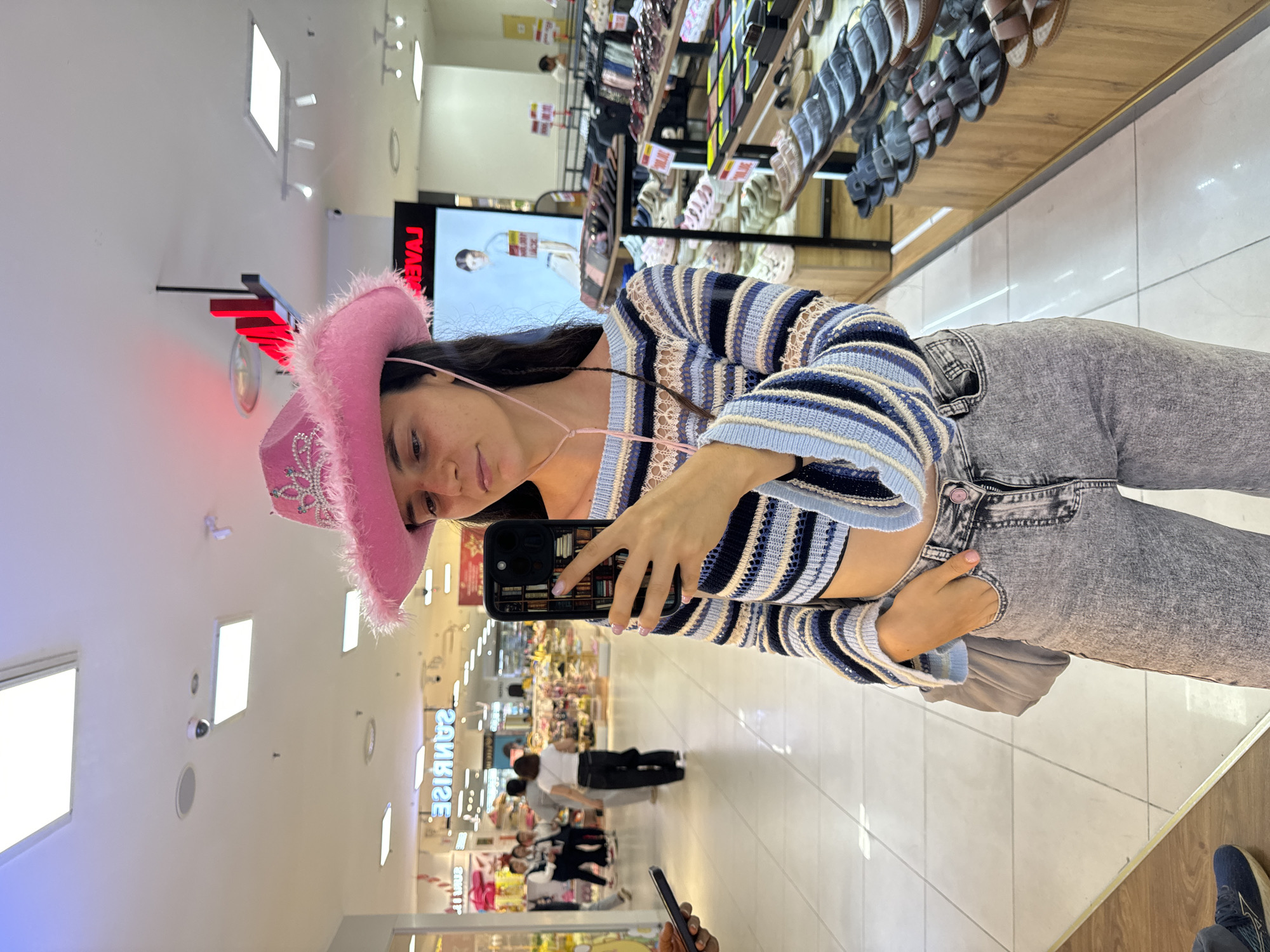 A young woman with dark hair wearing a pink cowboy hat, a blue and white striped cropped sweater, and gray jeans takes a selfie in front of a mirror. The reflection shows a brightly lit indoor space, likely a shopping mall or department store in Phú Thủy, Vietnam. Several people are visible in the background of the reflection. Shelves of shoes are visible behind her. A display screen is also visible behind the woman.