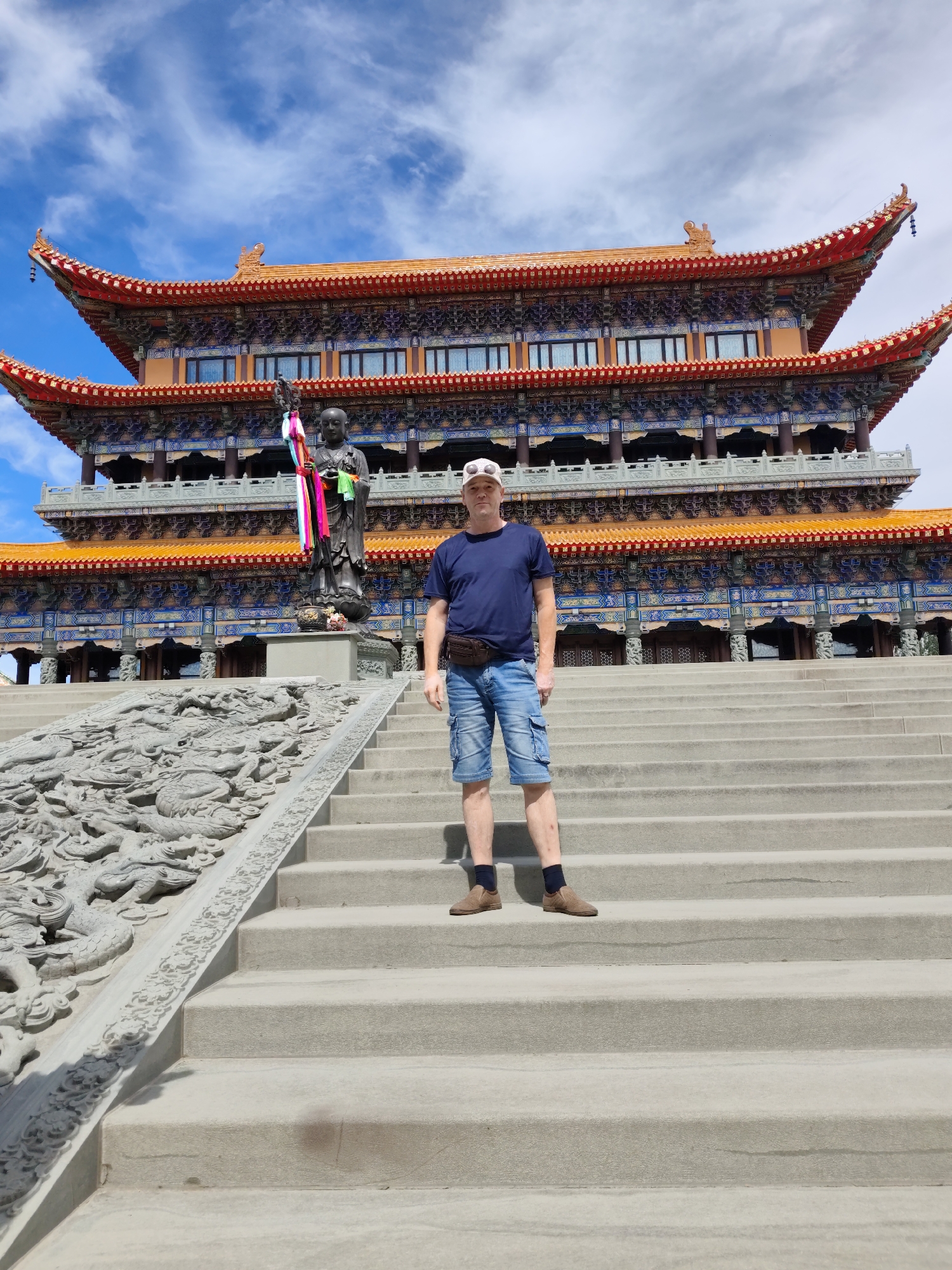 A man stands on a wide, outdoor concrete staircase in front of a large, ornate building with traditional East Asian architecture, identified as the Daqing Longze Temple in Daqing, China. The man, who appears to be middle-aged and Caucasian, is wearing a dark blue t-shirt, denim shorts, a brown fanny pack around his waist, and a light-colored baseball cap. He is positioned in the center of the frame, slightly to the right, with his body facing forward. Behind him and to his left, a dark bronze statue of a young monk holding a staff adorned with colorful ribbons stands on a pedestal. The temple building features multiple tiers with orange-tiled roofs, intricately decorated facades with blue and red accents, and rows of windows. The staircase itself is flanked on the left by a grey stone railing intricately carved with dragon motifs. The sky above is blue with scattered white clouds, suggesting daytime. The overall scene depicts a tourist at a significant cultural and religious site.