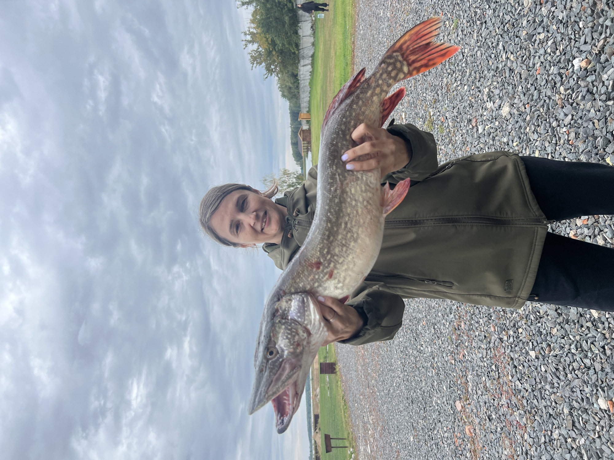 A woman stands outdoors on a gravelly surface, holding a large, freshly caught pike. She is wearing a dark green jacket and black pants. The pike, which is silver and green with red fins, is held horizontally in front of her, its mouth open. In the background, there is a body of water, a grassy field, trees, and some small buildings. The sky is overcast. There is a person in the far background on the right side of the image. The location is identified as Novo-Pichugo, Russia.