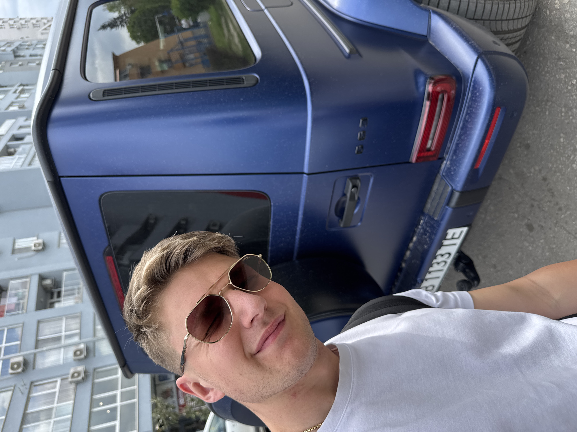 An outdoor, urban scene in Plovdiv, Bulgaria, depicts a young man smiling subtly at the camera, likely taking a selfie, with a luxury SUV behind him. The man has short, light-colored hair, is clean-shaven with light stubble, and wears a plain white crew-neck T-shirt with a thin gold chain. He is wearing aviator-style sunglasses with gold frames, which bear a 'CARRERA' logo on the temple.
Behind the man, a large, dark blue SUV, resembling a Mercedes-Benz G-Wagen with a matte or satin finish, is parked on what appears to be asphalt. The vehicle is visibly dusty and features distinctive rectangular LED taillights, a rear-mounted spare tire, and three vertically stacked characters on the right side of the rear door, likely part of its model badging. A partially visible Bulgarian license plate on the car reads 'B 1133 TE'.
In the background, a multi-story building with numerous windows and several exterior air conditioning units is visible, its reflection also appearing in the car's rear windows. The scene is brightly lit by daylight, suggesting a clear or partly cloudy day, and the man's relaxed smile conveys a positive mood.