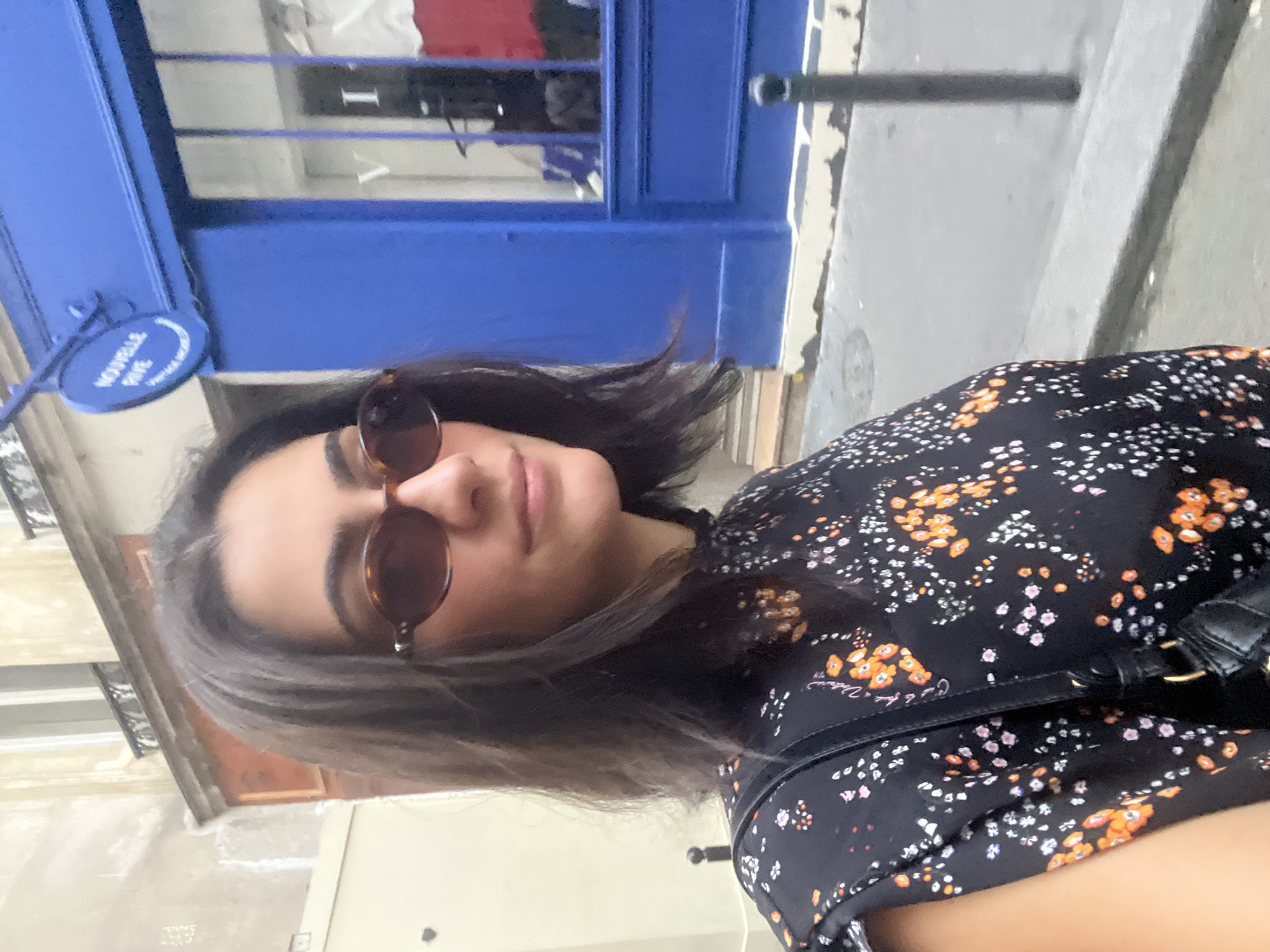 A medium shot captures a woman in Paris, France. She is facing the camera with her head turned slightly to the right, her face obscured by tortoiseshell sunglasses. Her dark, shoulder-length hair is slightly windblown. She is wearing a black dress with a floral pattern of orange, pink, and white flowers. A black purse with a strap is slung over her left shoulder.
Behind her, a bright blue building is visible with large glass windows. A circular sign with white lettering that reads "NOUVELLE RIVE" is mounted above the entrance. The street in front of the building is paved and there is a grey sidewalk. A black bollard stands on the sidewalk. The architecture suggests an urban setting in Paris.