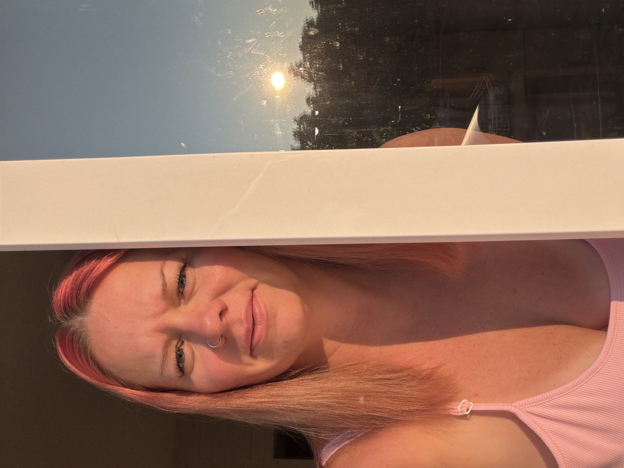 The image shows a woman with pink-tinted hair looking towards the camera. She is wearing a pink tank top and has a nose ring. The woman is positioned to the left of a white vertical frame, which partially obscures her body. To the right of the frame is a window that reflects the setting sun and trees. The lighting suggests it is either sunrise or sunset. The scene does not provide any specific visual cues to identify its location as Chaikovsky, Russia.