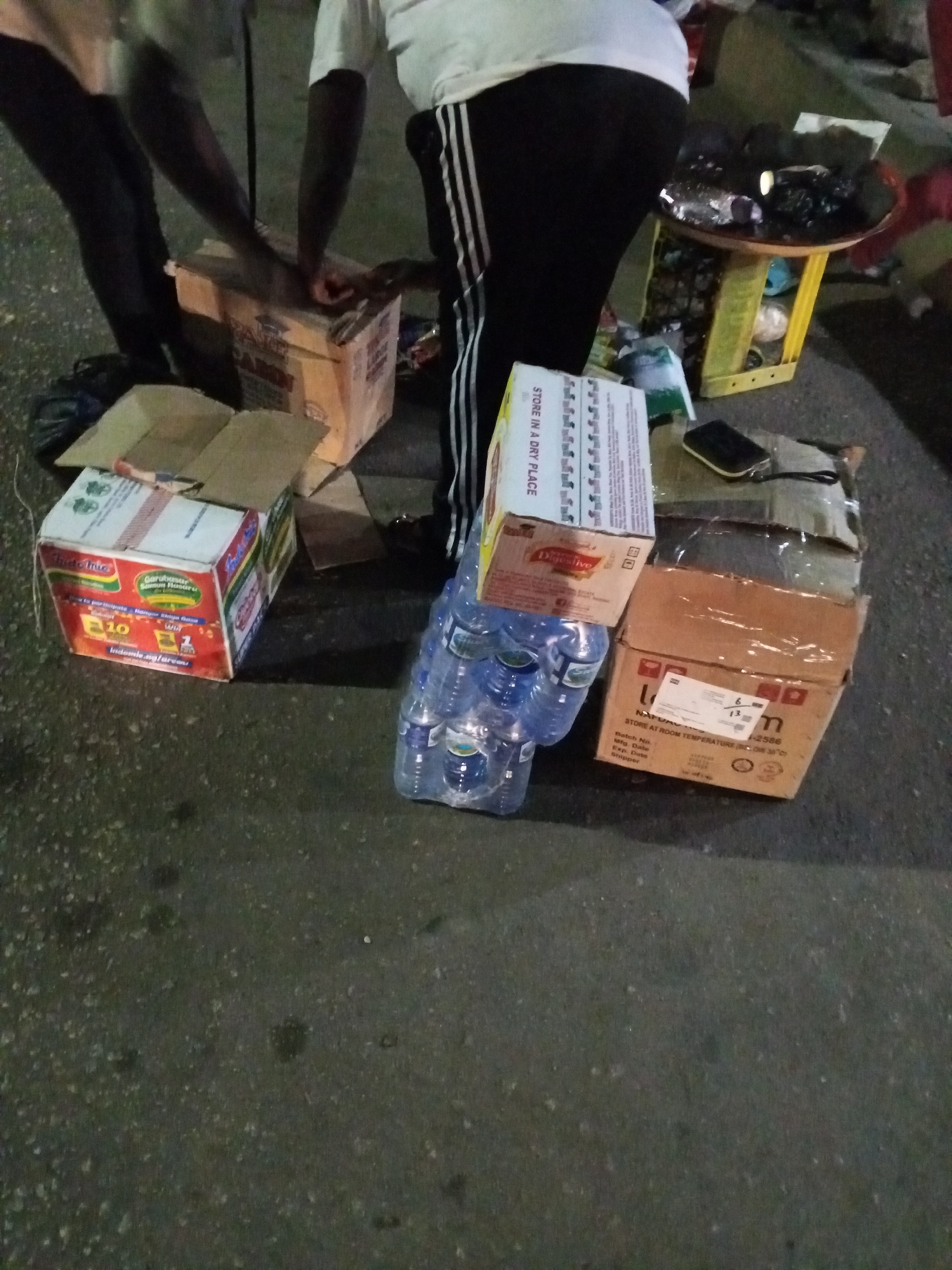In a nighttime scene in Bukuru, Nigeria, two individuals are near several boxes and items. One person wears dark pants and a lighter-colored shirt. The second person is wearing black pants with white stripes down the side and a white shirt. There are several boxes. One box is labeled "Indomie" and features images of noodles and promotional text. Another box has "STORE IN A DRY PLACE" printed on it. A stack of bottled water is also visible. A small yellow table with items on top is located behind the individuals and boxes.